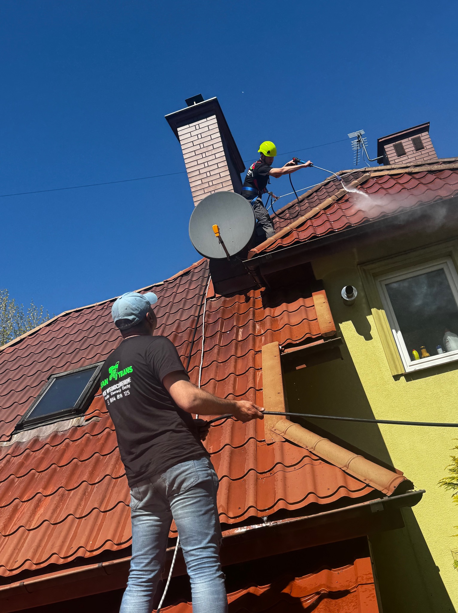Dwóch mężczyzn myje dach domu z czerwonej dachówki za pomocą myjki ciśnieniowej. Jeden stoi na dachu, drugi podaje wąż. Widoczne zabrudzenia i czysta powierzchnia.
