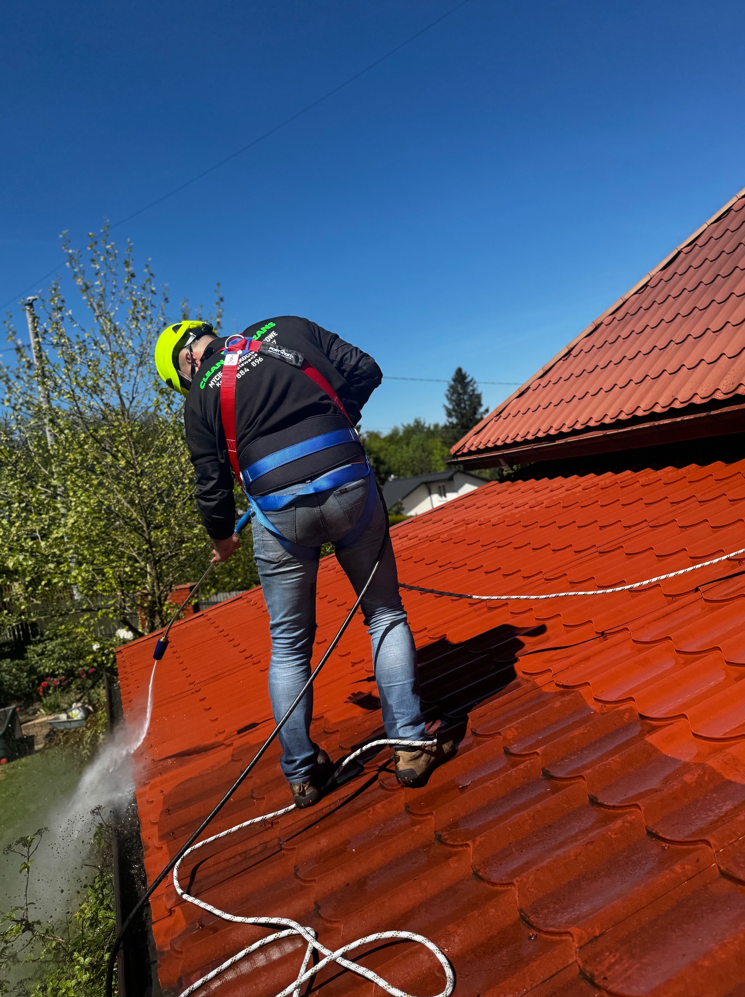 Pracownik w kasku i uprzęży myje czerwoną dachówkę z użyciem myjki ciśnieniowej. Widoczne strumienie wody i czyszczona powierzchnia dachu. Błękitne niebo w tle.