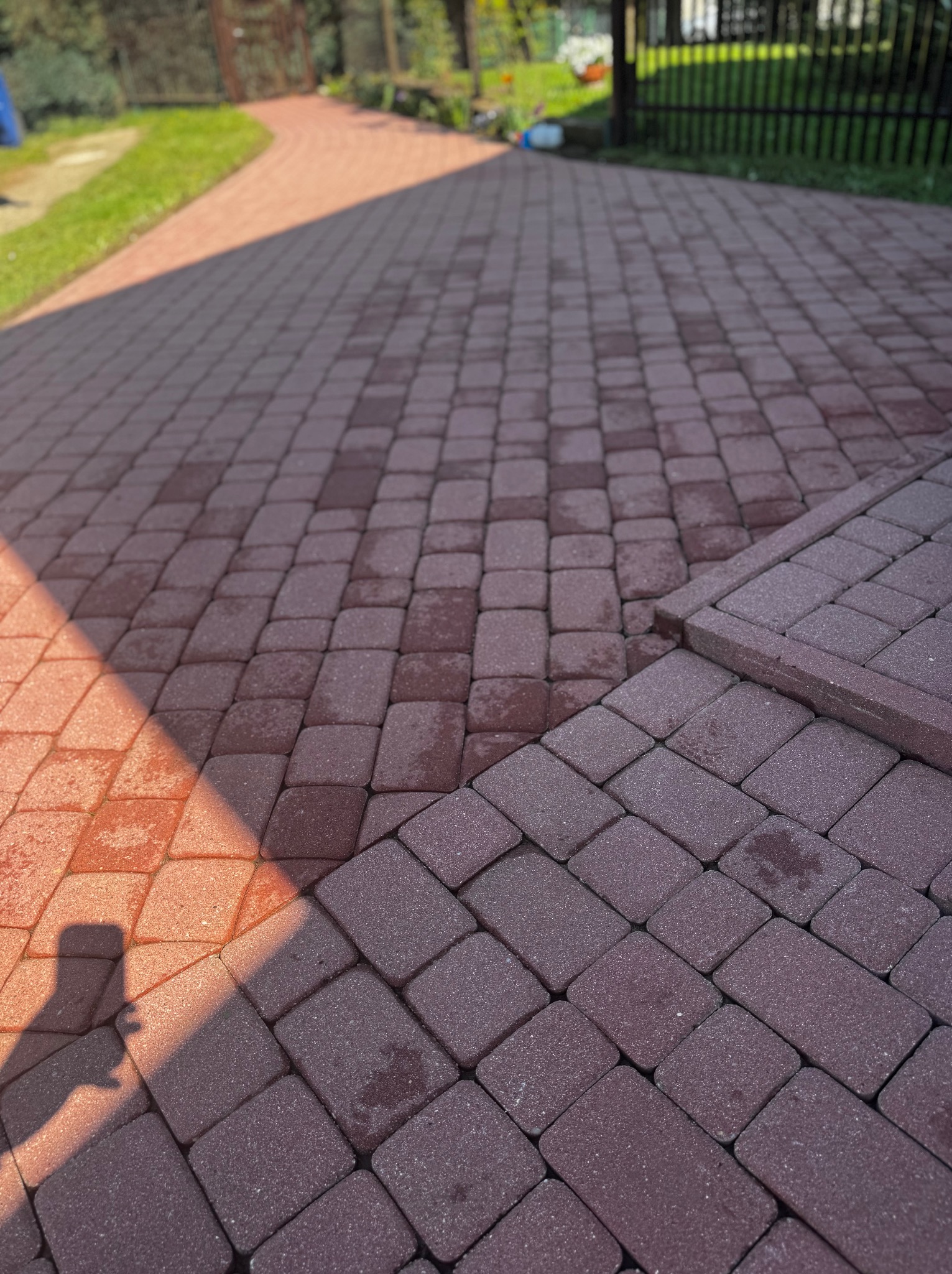 Czerwona kostka brukowa ułożona w jodełkę, częściowo mokra i zacieniona, z widocznym cieniem osoby. Fragment ścieżki i ogrodzenie w tle.