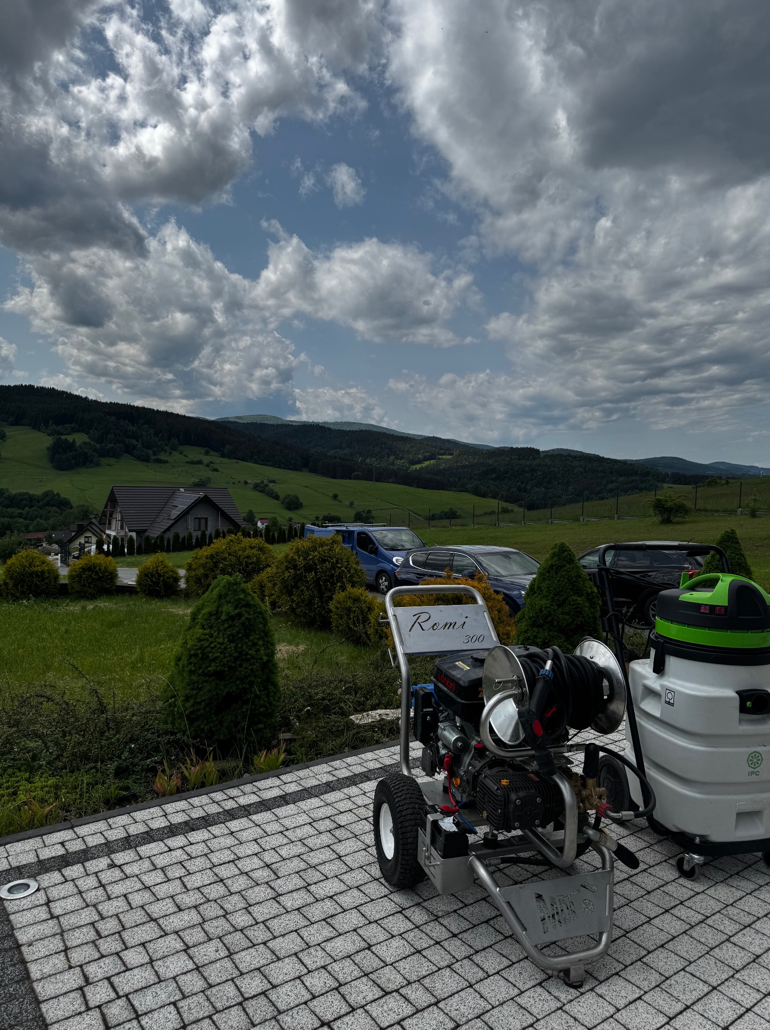 Profesjonalna myjka ciśnieniowa 'Remi 300' na kostce brukowej, z odkurzaczem przemysłowym w tle, na tle malowniczego krajobrazu z domem i wzgórzami.