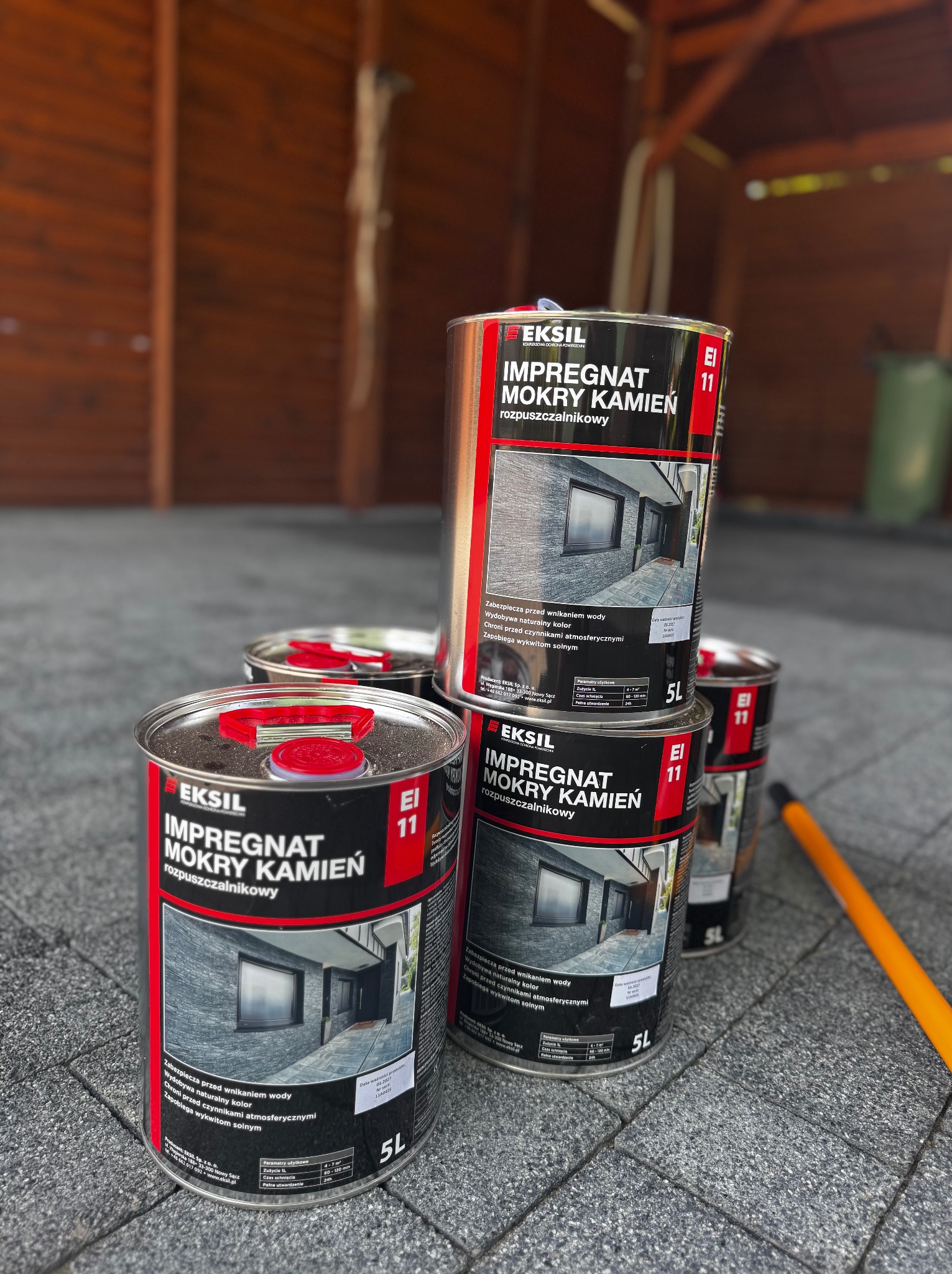 Puszki z impregnatem do kamienia 'Eksil' na szarej kostce brukowej, z drewnianym tłem. Widoczne etykiety z opisem, zabezpieczające przed wodą i czynnikami atmosferycznymi.