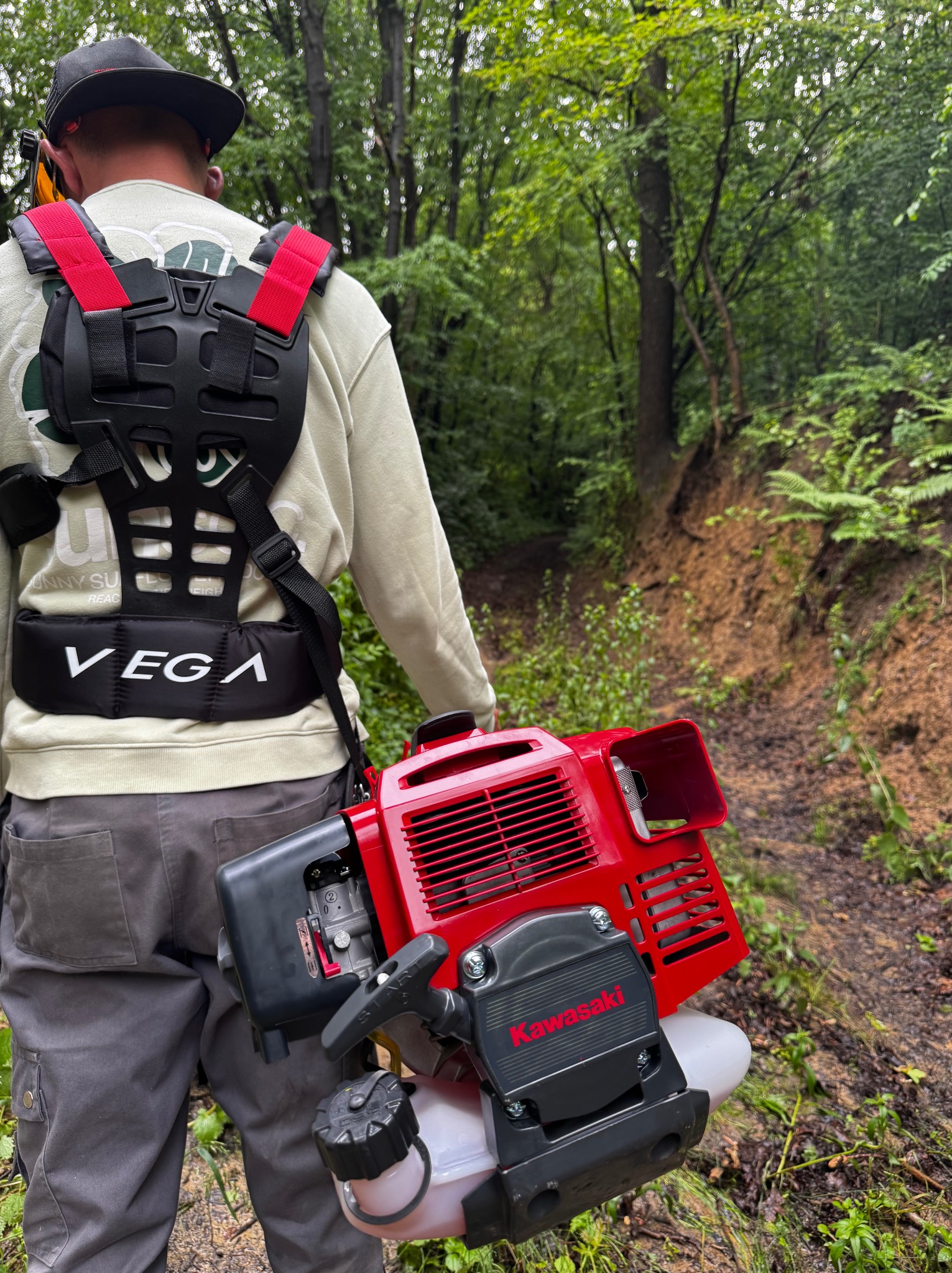 Mężczyzna z szelkami i kosą spalinową Kawasaki na tle zalesionego terenu. Widoczna ścieżka i naturalne otoczenie, sugerujące prace porządkowe w terenie.
