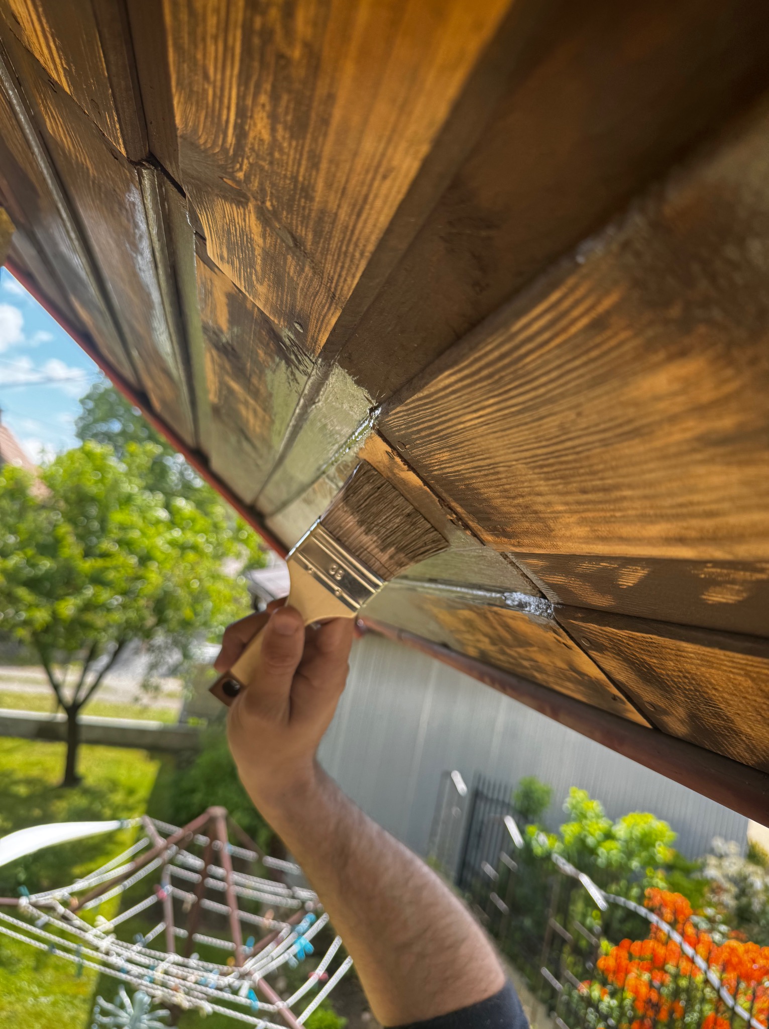 Malowanie drewnianej podbitki dachowej pędzlem, widok z dołu na tle zieleni. Prace renowacyjne z bliska, podkreślona faktura drewna i detale malowania.