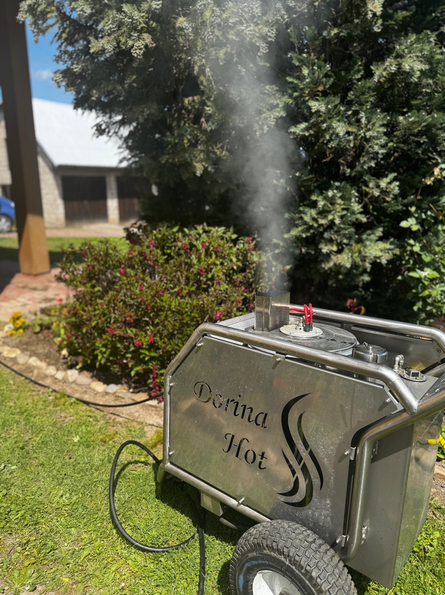 Generator pary 'Derina Hot' na trawniku, z dyszy unosi się para. W tle budynek gospodarczy i zieleń. Urządzenie stoi na kołach, gotowe do użycia.
