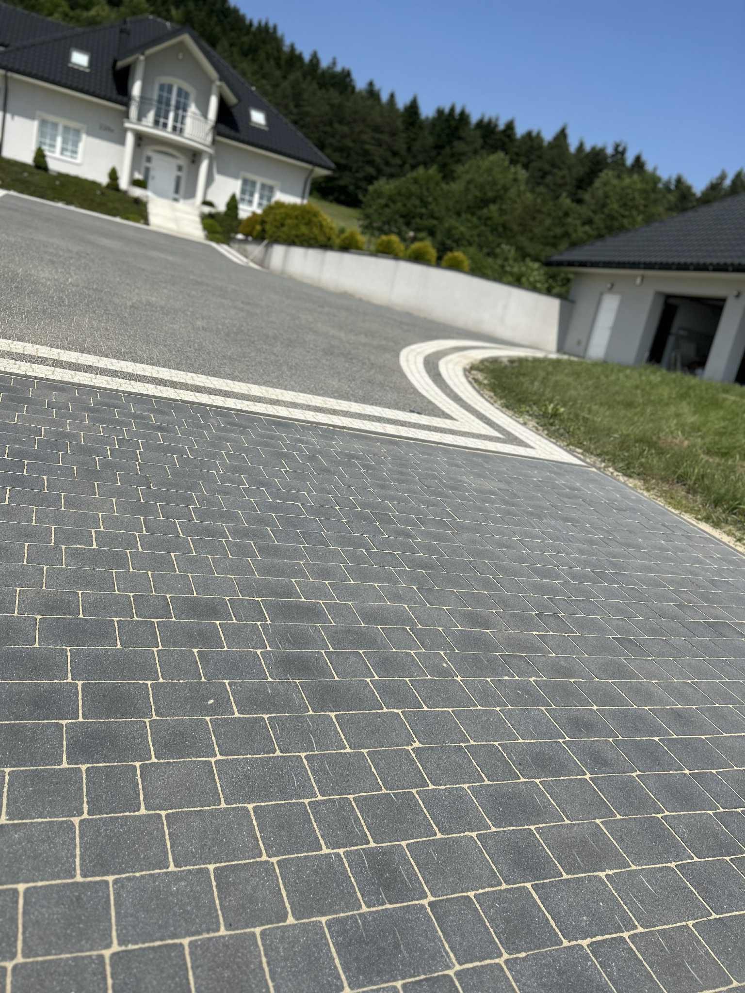 Ułożona, szara kostka brukowa na podjeździe do domu z białymi elementami dekoracyjnymi i zielenią w tle, w słoneczny dzień. Widok z perspektywy chodnika.