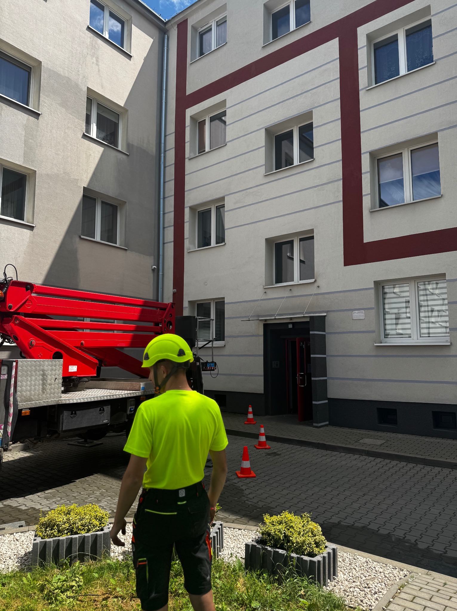 Pracownik w kasku i jaskrawej koszulce stoi przed budynkiem, obok podnośnika koszowego i pachołków, prawdopodobnie przygotowując się do mycia elewacji.