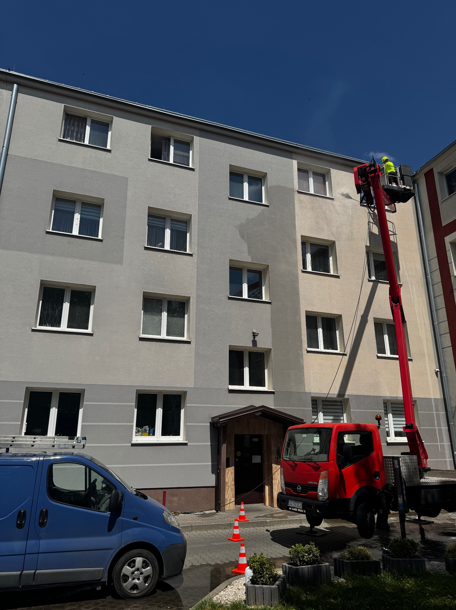 Pracownik w żółtym kombinezonie myje elewację budynku z podnośnika koszowego. Widoczny niebieski bus i czerwona ciężarówka. Na ziemi stoją pachołki.