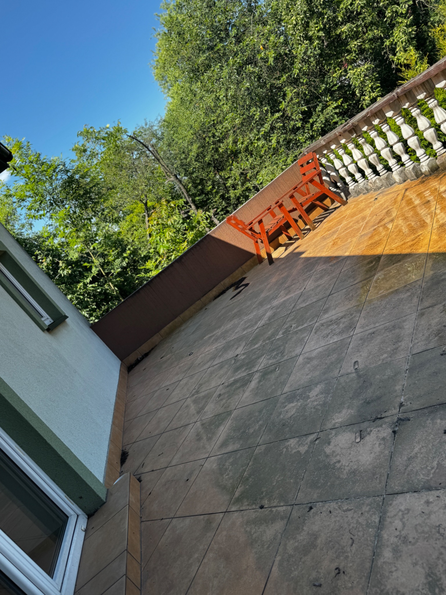 Ukośne ujęcie zabrudzonego balkonu z terakotą, z widocznymi plamami i osadami. W tle zieleń drzew i błękit nieba. Na balkonie stoi pomarańczowe krzesło.