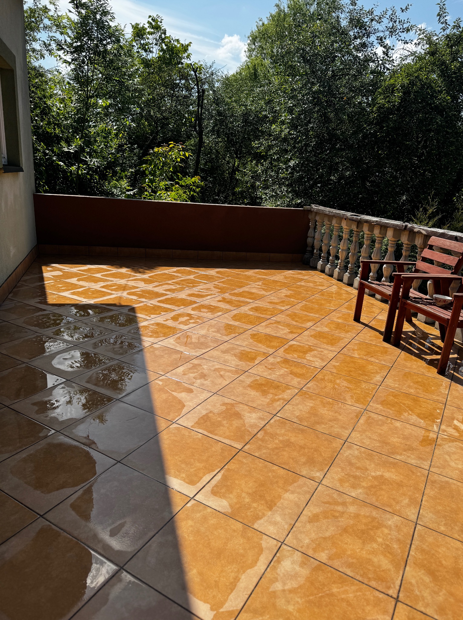 Mokry, pomarańczowy taras z płytkami po umyciu, z widokiem na zieleń drzew. Dwa drewniane krzesła stoją z boku, a na płytkach odbija się niebo.