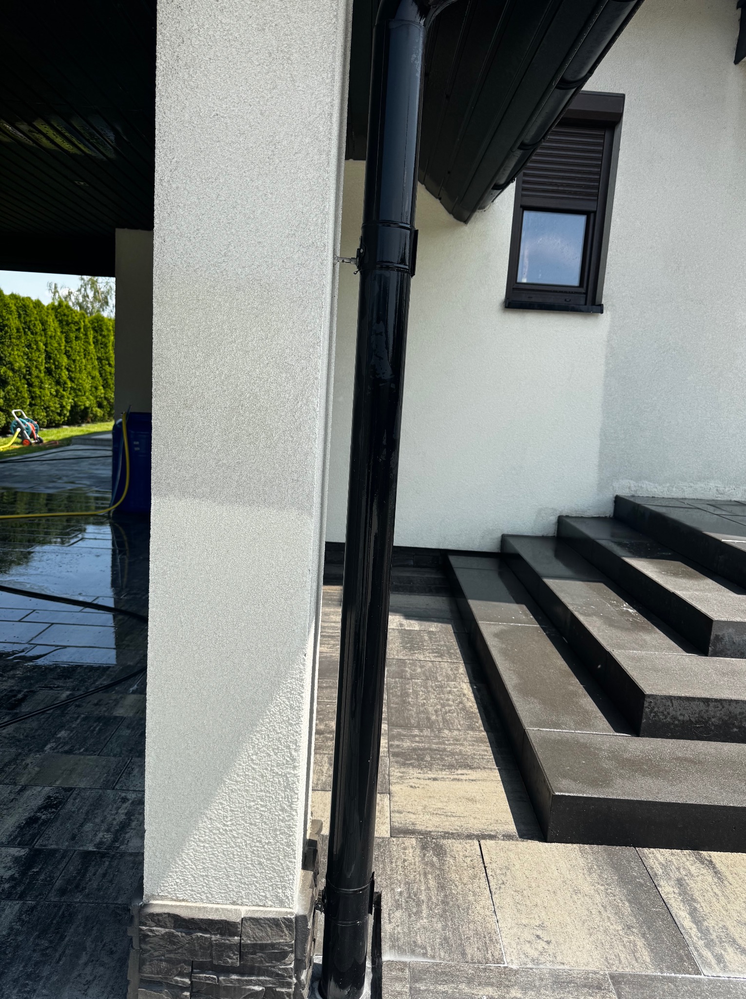 Świeżo umyta elewacja domu z czarną rynną i schodami z kostki brukowej. Widoczne mokre powierzchnie i sprzęt do mycia ciśnieniowego w tle.