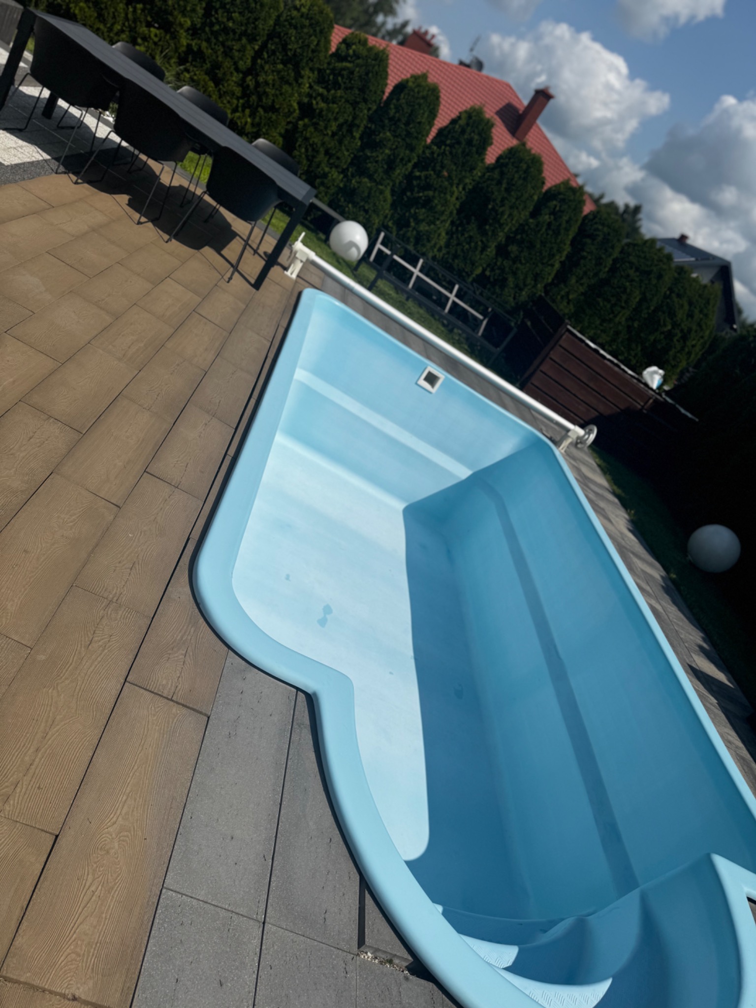 Niebieski basen z kompozytowymi deskami tarasowymi i kostką brukową, otoczony zielenią. Widok z góry, z perspektywą na dom z czerwoną dachówką i błękitne niebo.