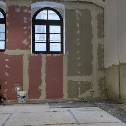 Kmk team sp zoo - Wnętrze w trakcie szpachlowania: ściany częściowo pokryte gładzią, widoczne okna z czarną ramą i wiadro z masą szpachlową. Podłoga zabezpieczona folią.