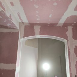 Kmk team sp zoo - Wnętrze w trakcie szpachlowania: różowe płyty gipsowe z białymi pasami szpachli na ścianach i suficie. Widoczne łukowe przejście. W tle lampa budowlana.
