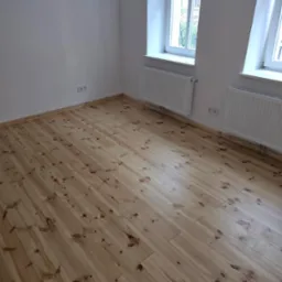 Jasne, drewniane panele podłogowe w pustym pokoju z białymi ścianami, grzejnikami i dwoma oknami. Widok na miasto przez okna.