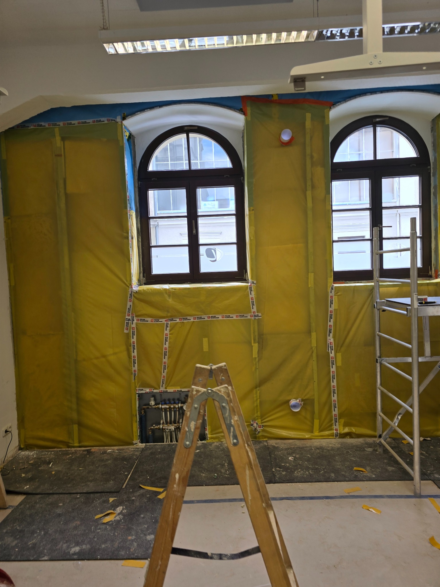 Przygotowanie do remontu: ściany zabezpieczone żółtą folią, widoczne okna łukowe, drabina drewniana i rusztowanie. Instalacja grzewcza odsłonięta.