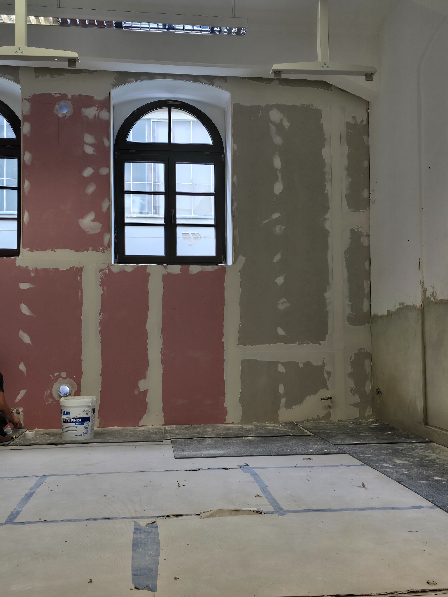 Wnętrze w trakcie szpachlowania: ściany częściowo pokryte gładzią, widoczne okna z czarną ramą i wiadro z masą szpachlową. Podłoga zabezpieczona folią.