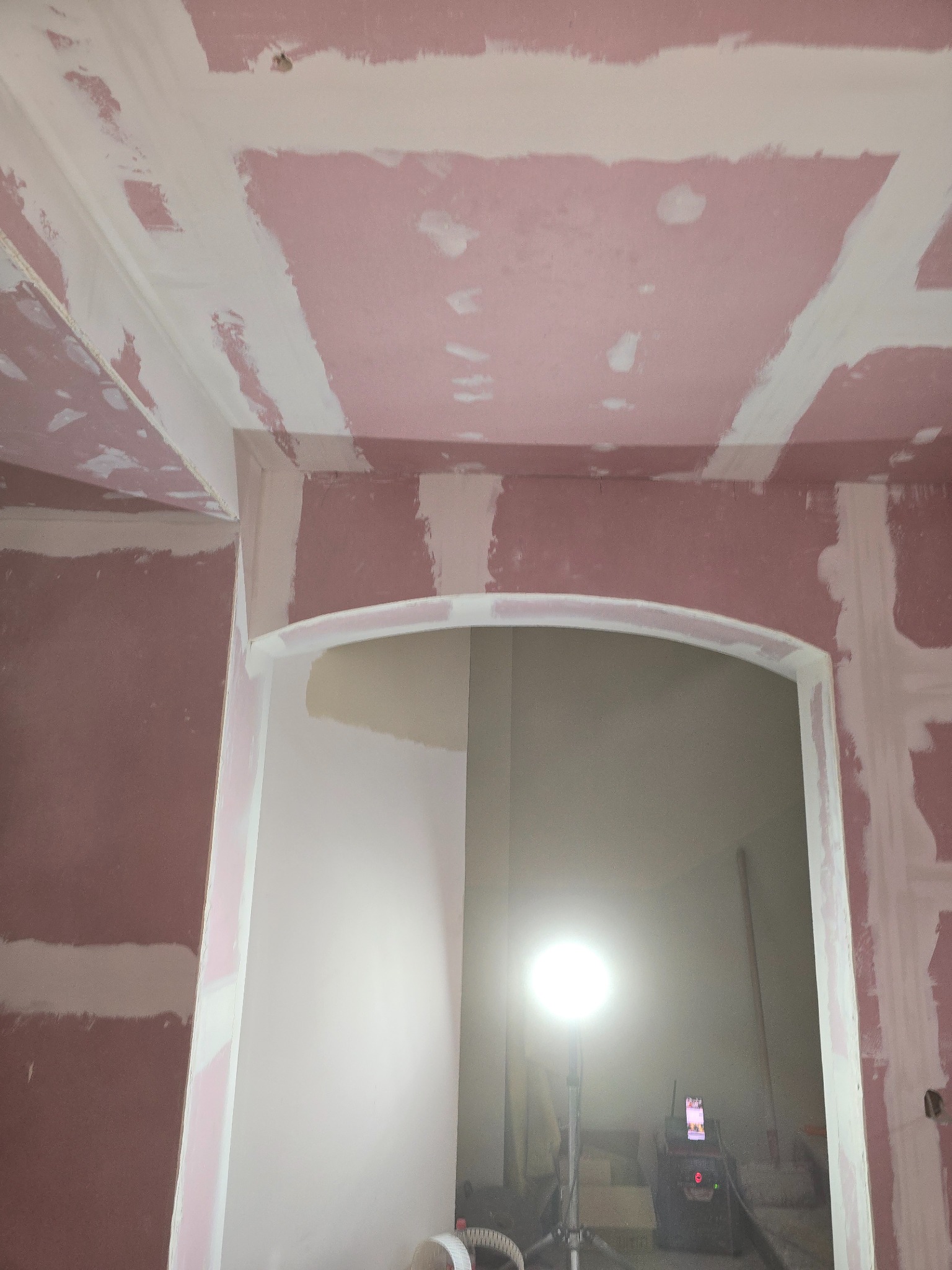 Wnętrze w trakcie szpachlowania: różowe płyty gipsowe z białymi pasami szpachli na ścianach i suficie. Widoczne łukowe przejście. W tle lampa budowlana.