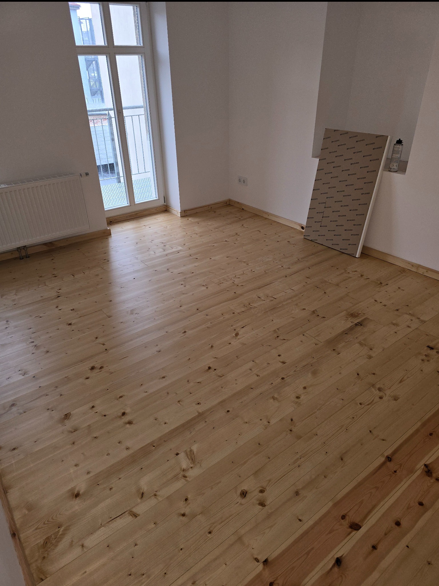 Jasne, drewniane panele podłogowe w pustym pokoju z oknem balkonowym i grzejnikiem. Przy ścianie leżą płyty zabezpieczające. Widok na gotową podłogę po ułożeniu.