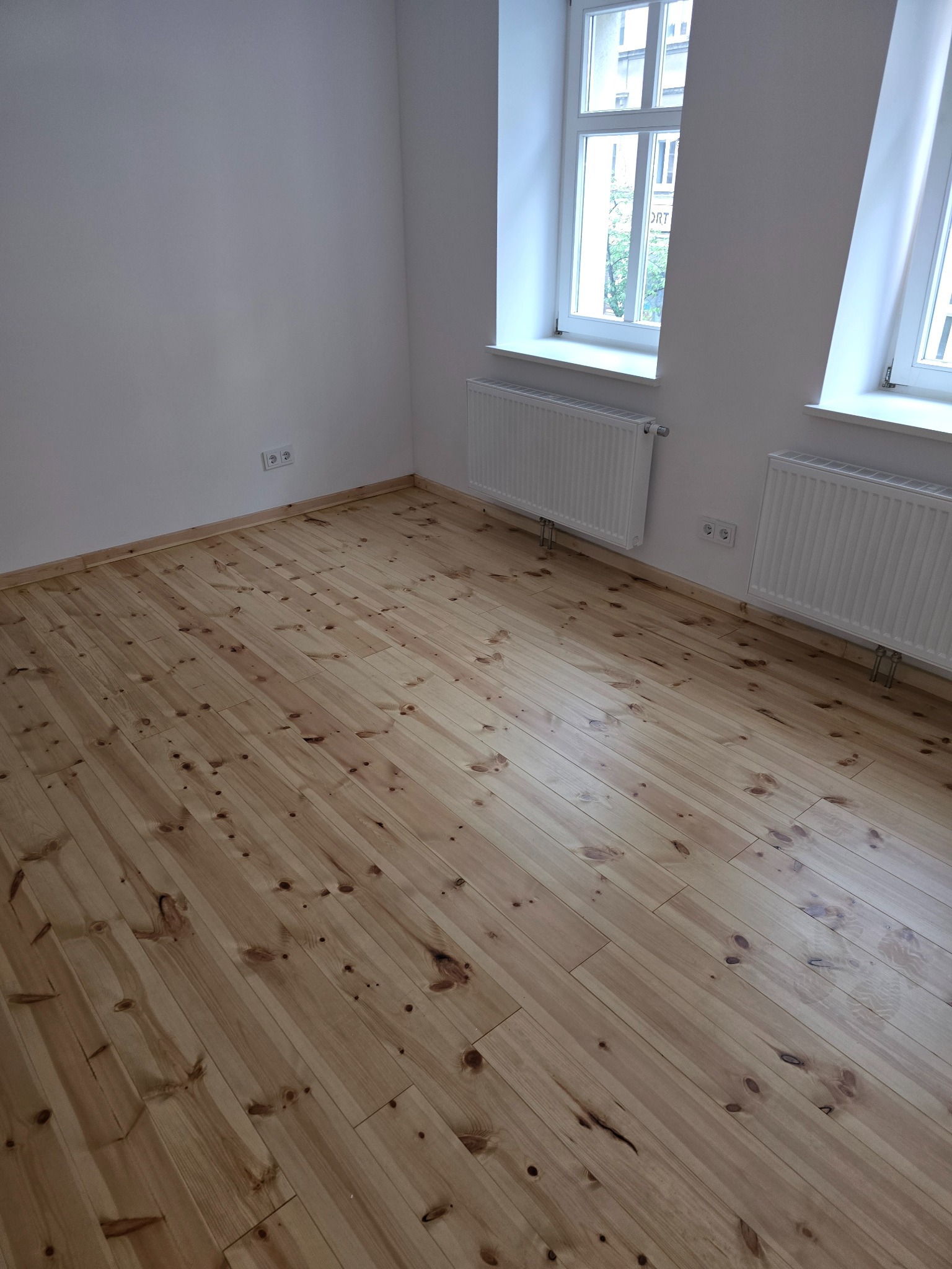 Jasne, drewniane panele podłogowe w pustym pokoju z białymi ścianami, grzejnikami i dwoma oknami. Widok na miasto przez okna.