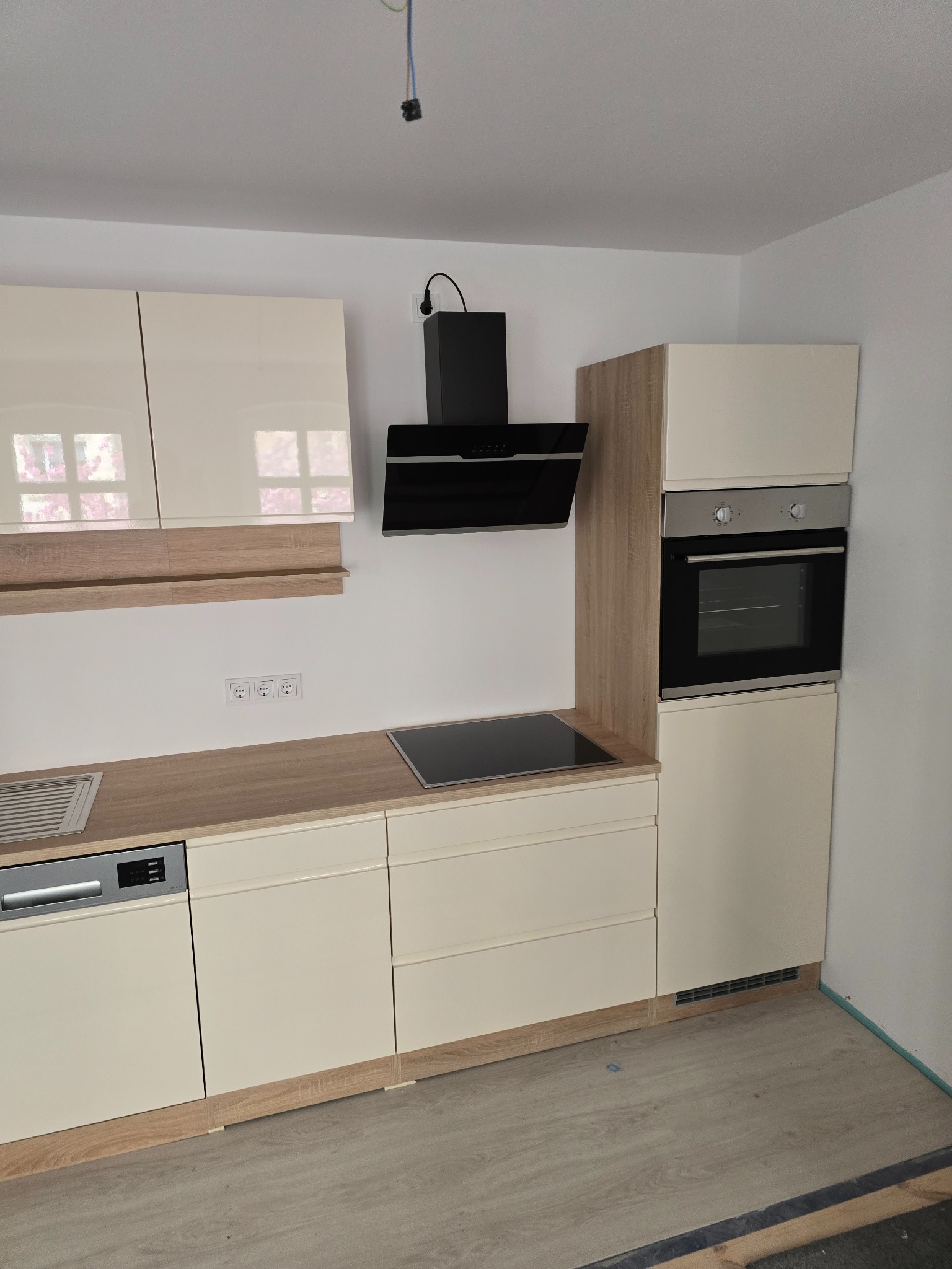 Jasna, nowoczesna zabudowa kuchenna z piekarnikiem, płytą indukcyjną i okapem. Beżowe fronty szafek, drewnopodobny blat i panele. Widoczne gniazdka elektryczne.