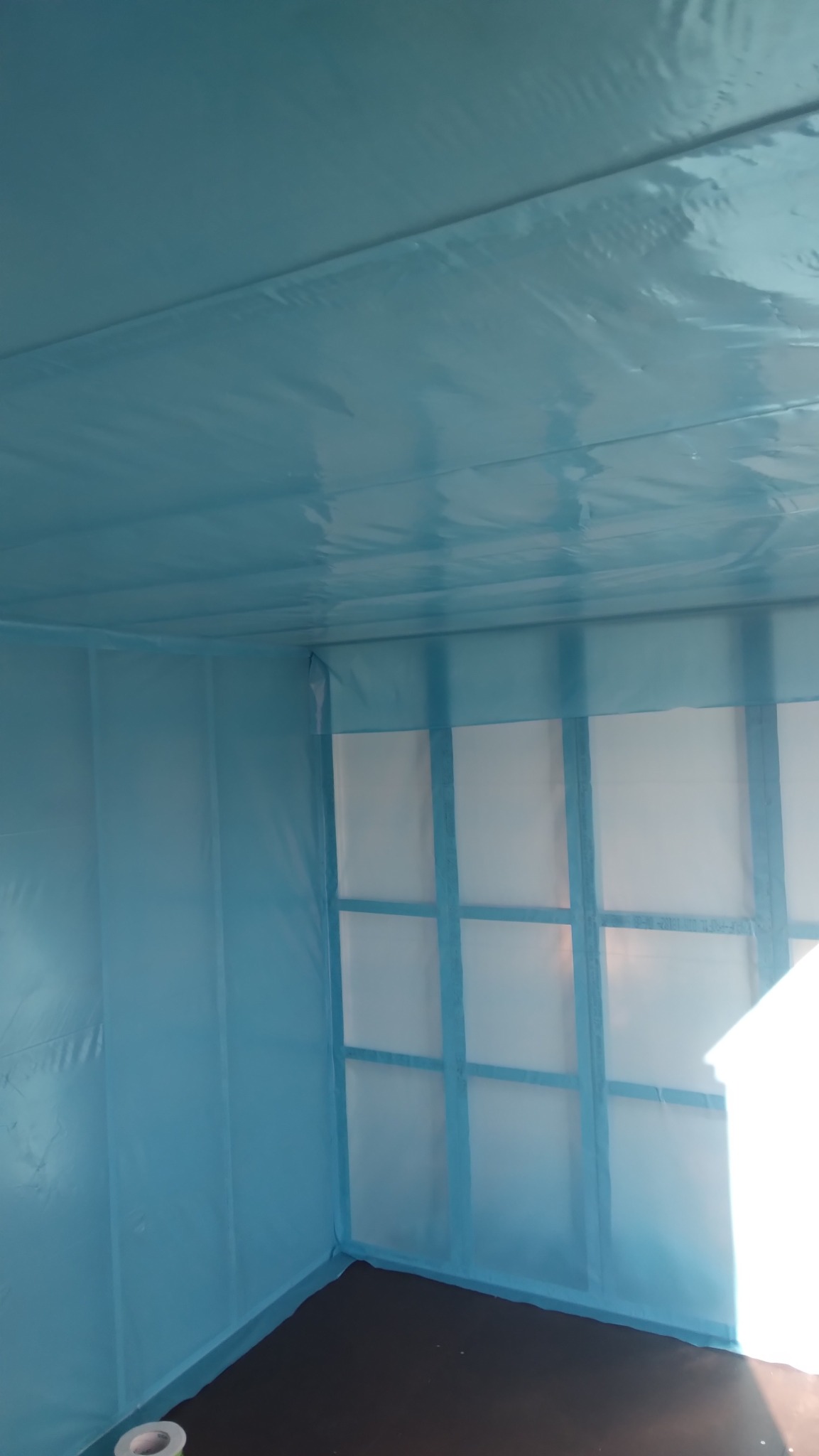 Pokój zabezpieczony folią i taśmą malarską przed remontem, widoczne ramy okienne i podłoga. Przygotowanie pomieszczenia do prac wykończeniowych, ochrona przed zabrudzeniami.