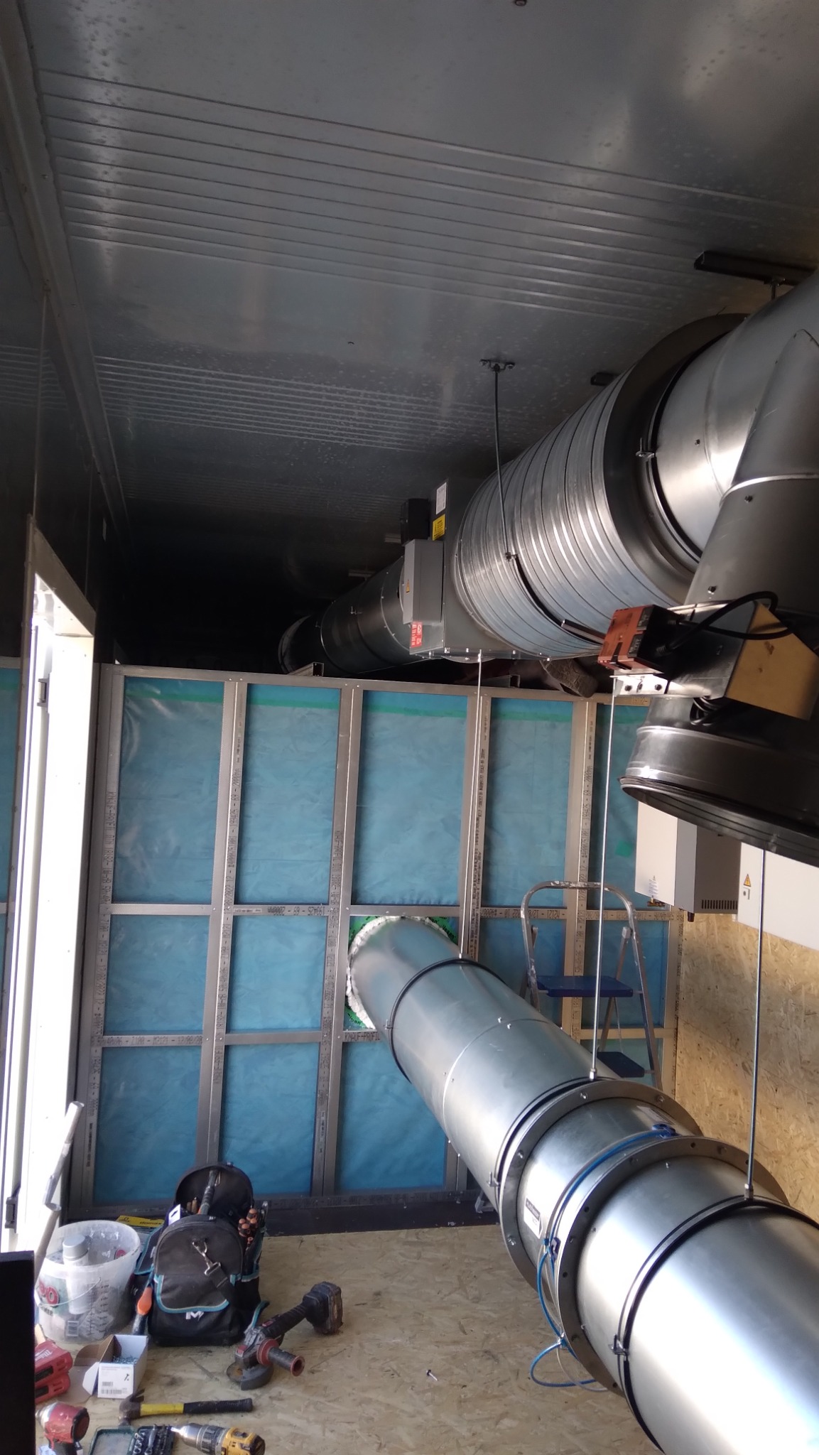 Montaż systemu wentylacji: metalowe rury, narzędzia i drabina w przestrzeni z niebieską izolacją. Widok z dołu, ukazujący etap instalacji.