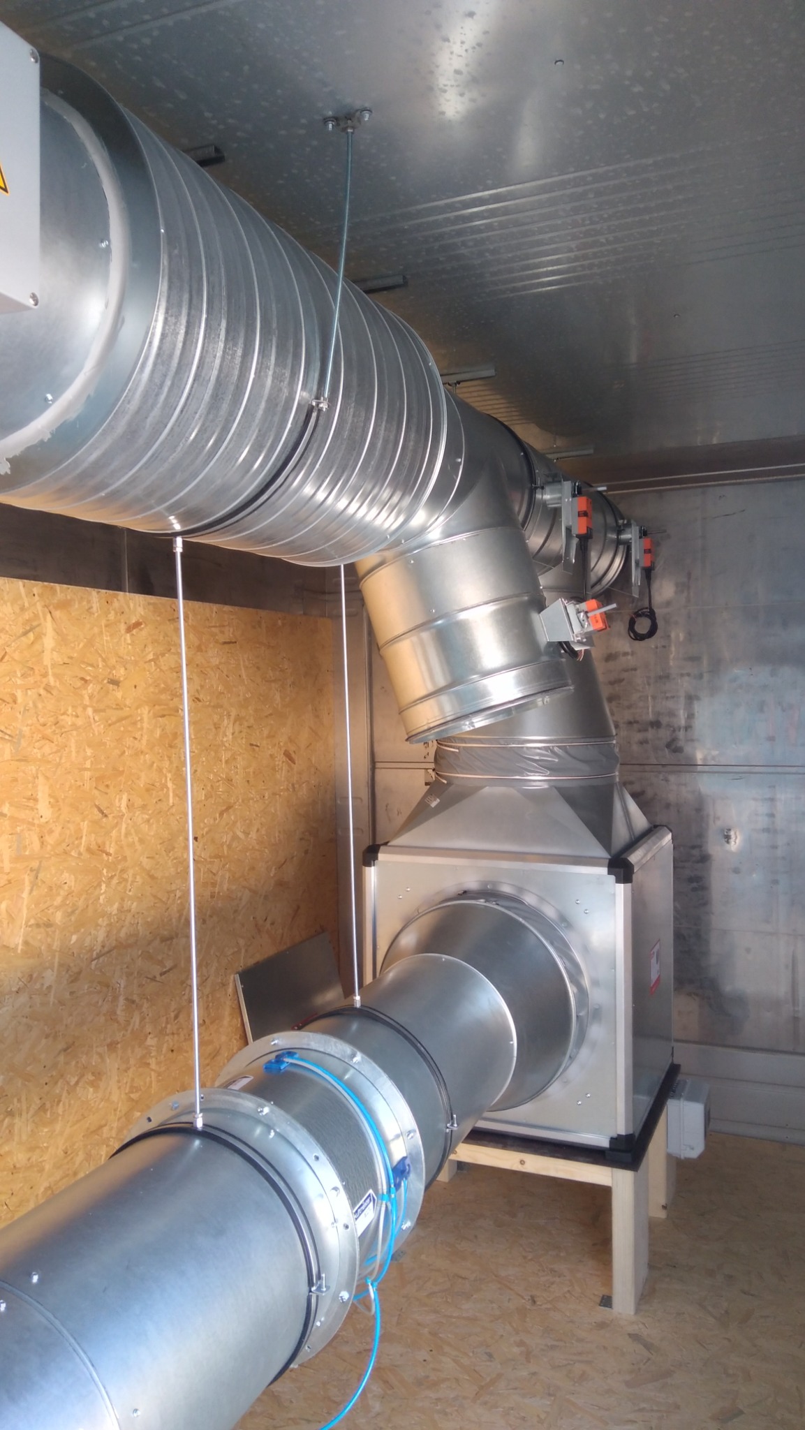 Metalowe rury wentylacyjne, połączone pod kątem, zamontowane w pomieszczeniu z panelami ściennymi. Widoczne elementy mocujące i przewody.