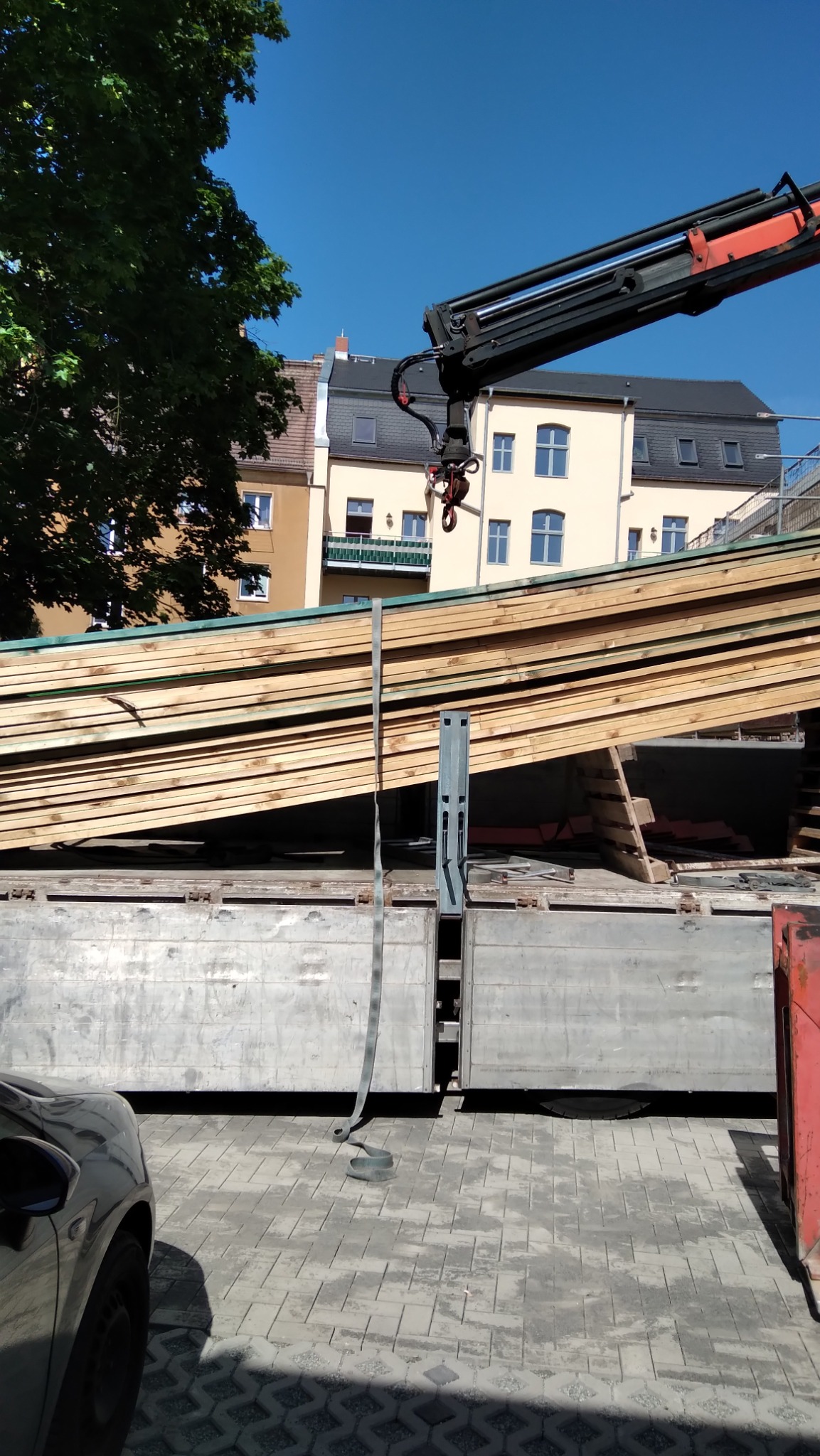 Transport desek na budowę. Żuraw unosi pakiet drewnianych desek na tle budynku mieszkalnego i błękitnego nieba w słoneczny dzień.