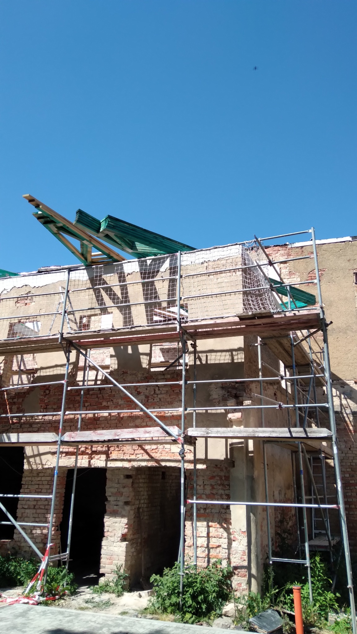 Fragment remontowanego budynku z cegły, z widocznymi rusztowaniami, drewnianymi elementami konstrukcyjnymi i fragmentami siatki zabezpieczającej, na tle błękitnego nieba.