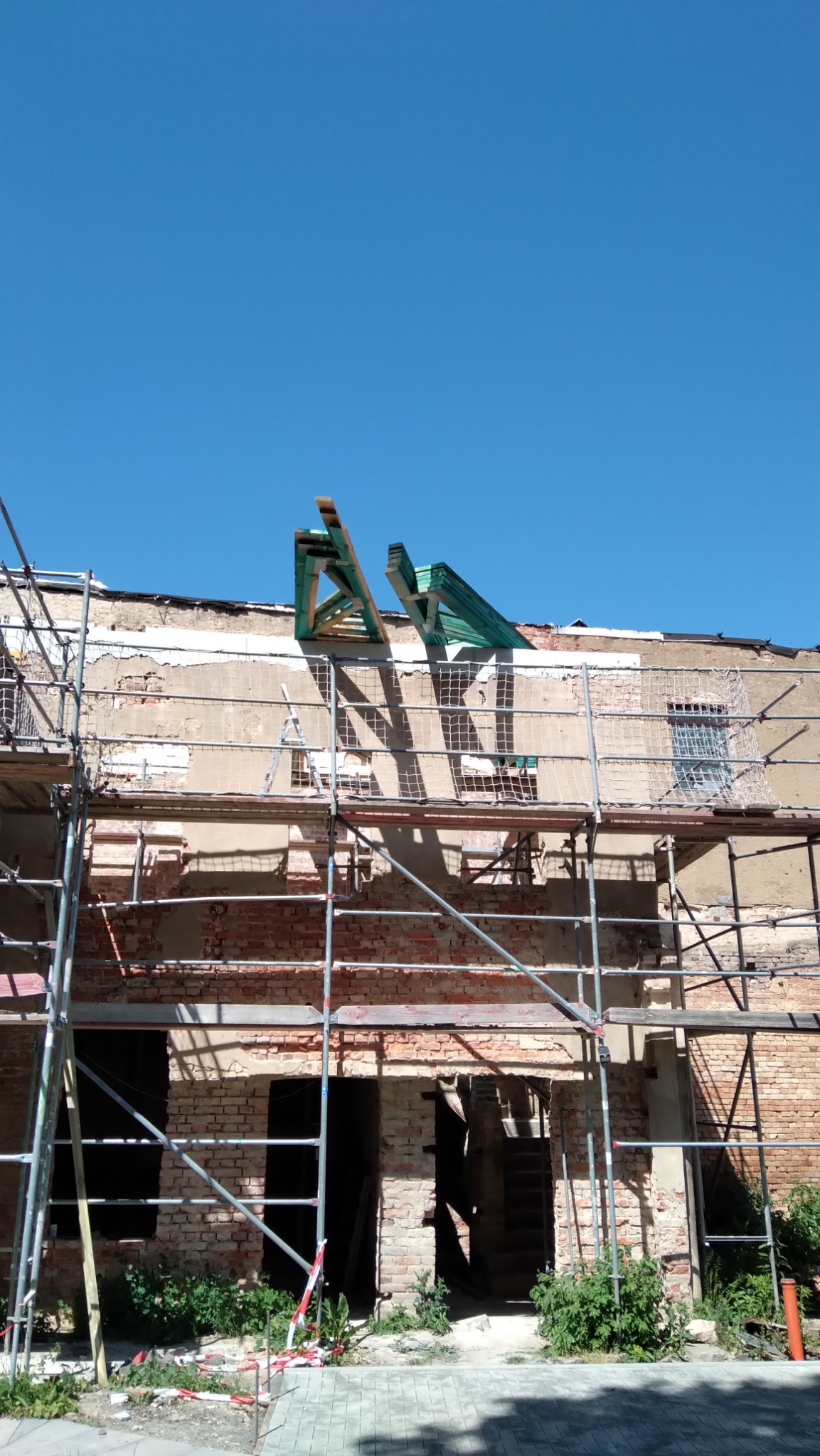 Budynek w trakcie remontu z odsłoniętą cegłą, rusztowaniami i elementami nowej konstrukcji dachu na tle błękitnego nieba.