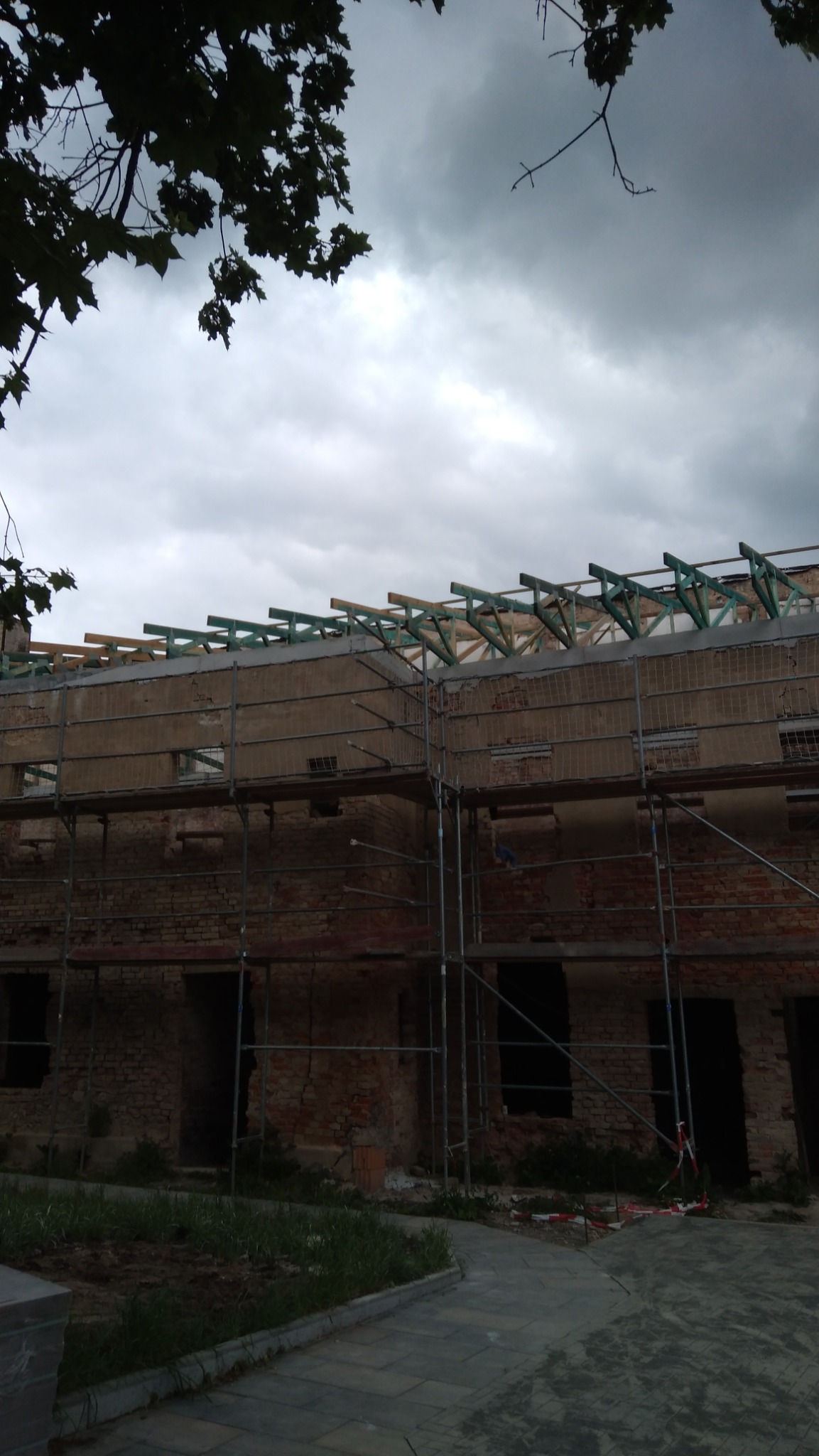 Budynek w trakcie remontu z rusztowaniem, widoczne fragmenty muru z cegły, elementy konstrukcji dachu i pochmurne niebo. Etap prac budowlanych.