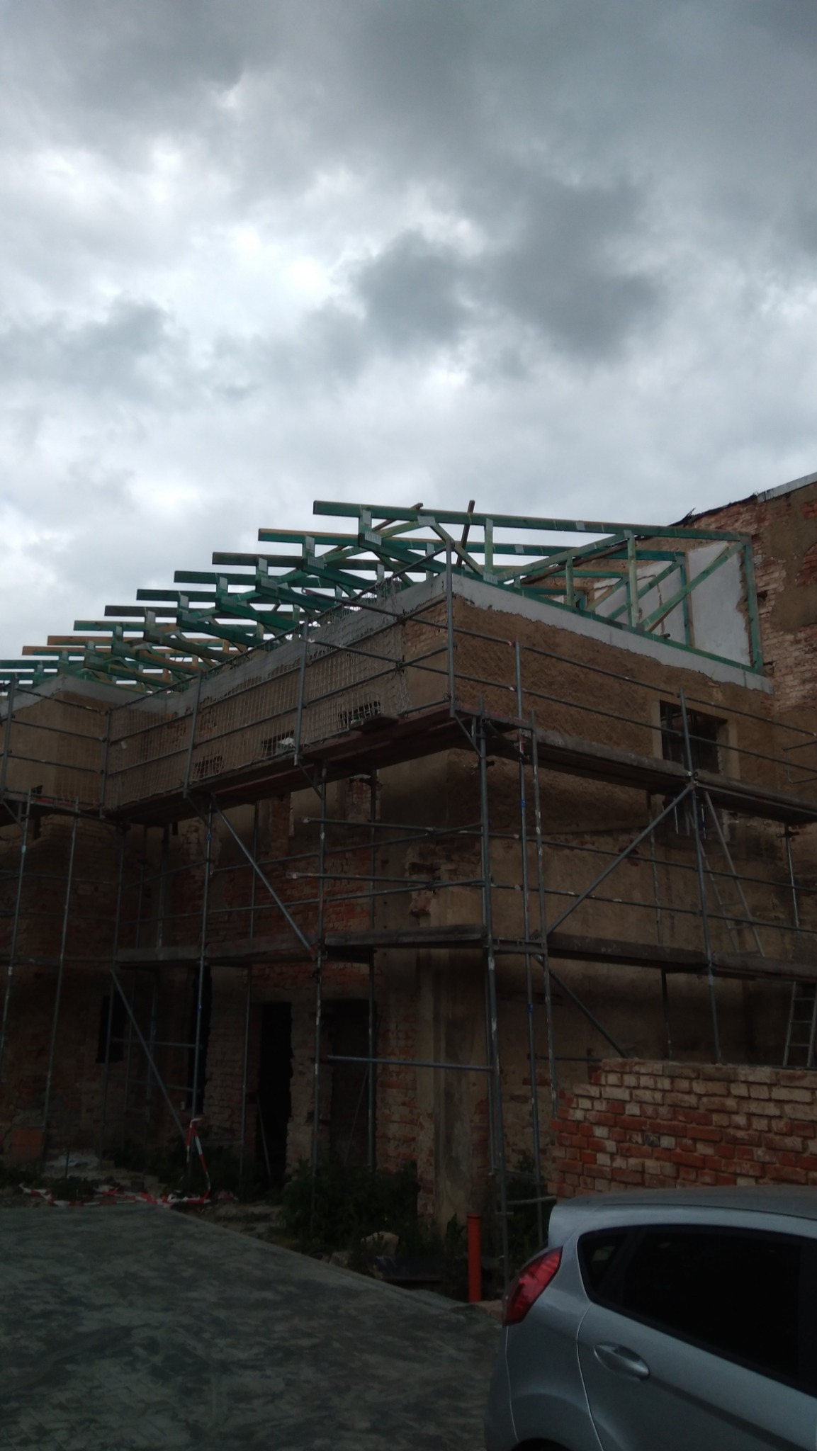 Renowacja budynku w toku: rusztowania, odsłonięta cegła, drewniana konstrukcja dachu. Szara, pochmurna pogoda. Fragment auta w rogu kadru.