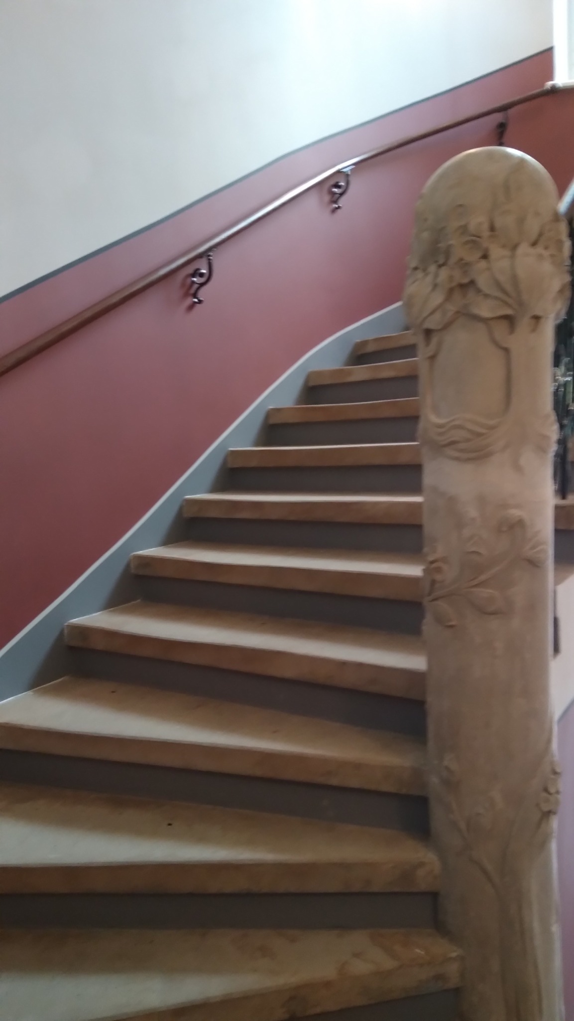 Zakręcone schody z drewnianymi stopniami i zdobioną, kamienną balustradą. Ściana w odcieniach czerwieni, brązowa poręcz. Widok z dołu, skupienie na detalach.