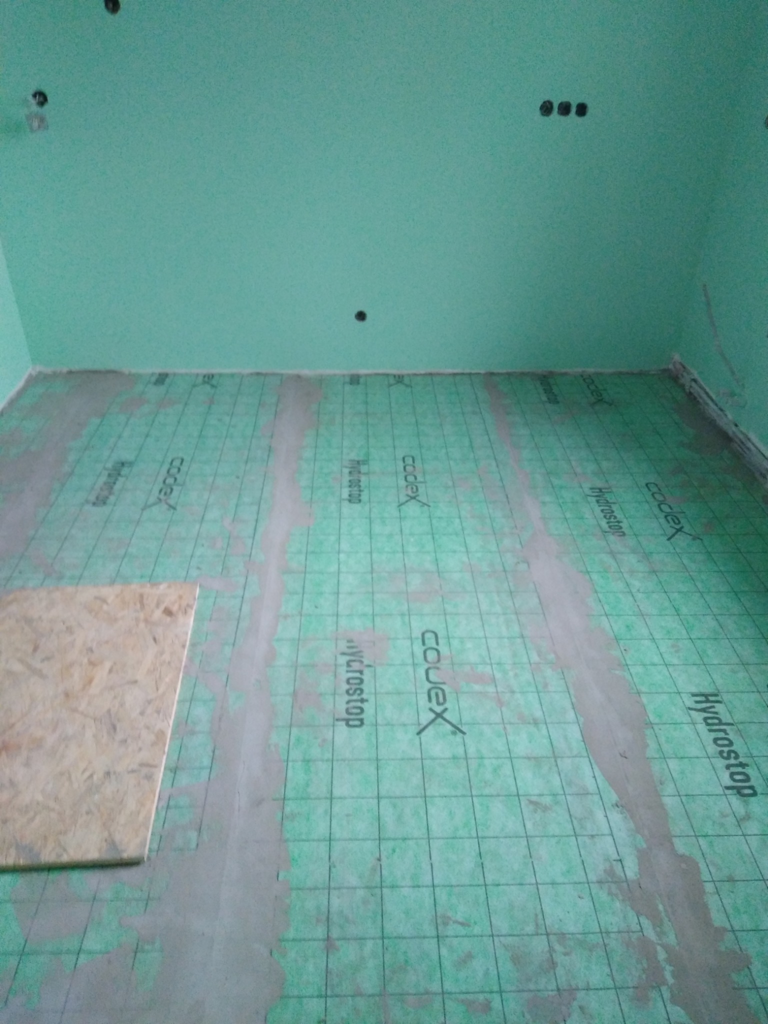 Podłoga w trakcie hydroizolacji, zielona mata z nadrukiem, częściowo pokryta wylewką. W rogu płyta OSB. Ściany pomalowane na zielono.