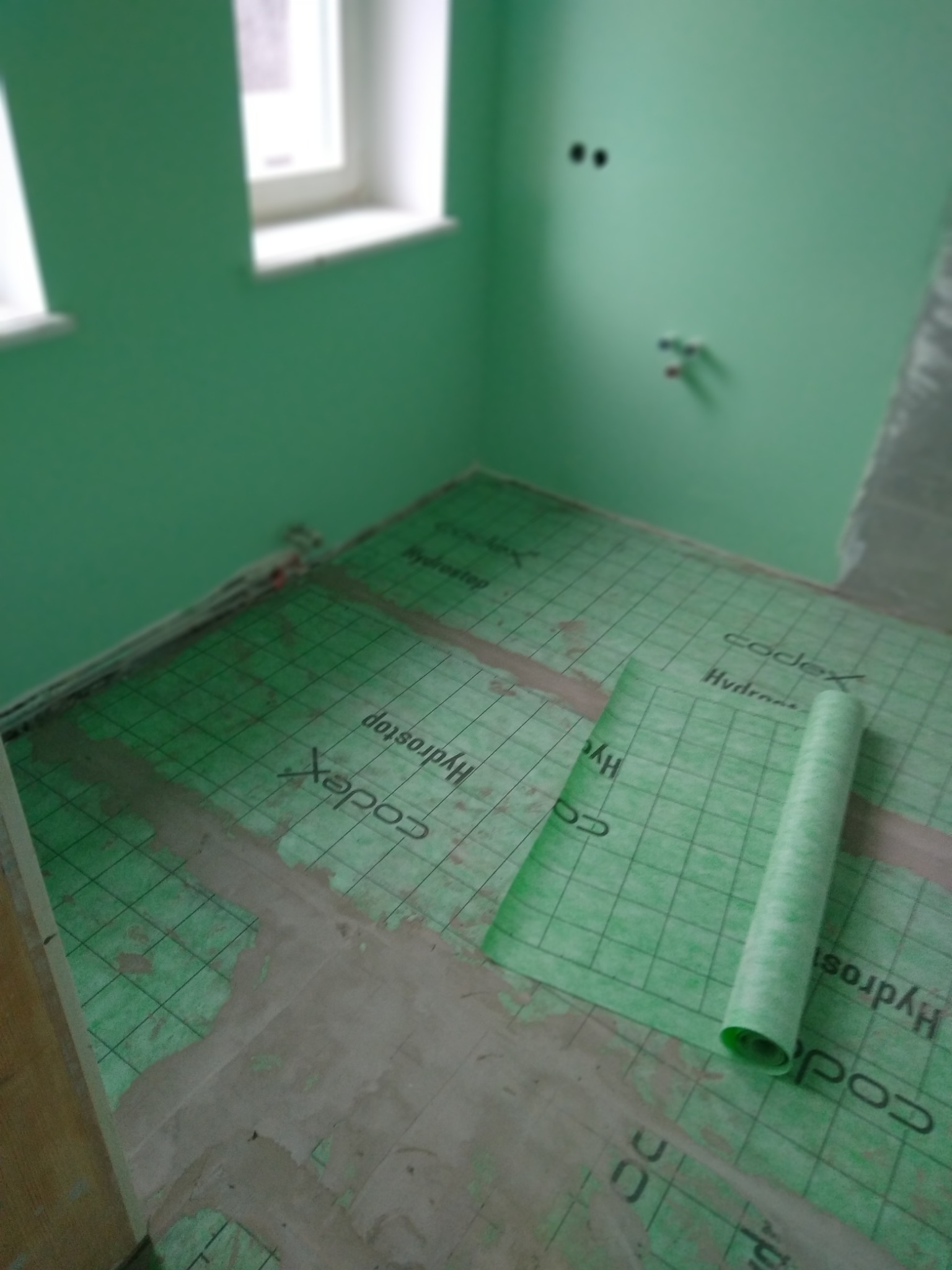 Wnętrze łazienki w trakcie prac wykończeniowych. Widoczna zielona mata hydroizolacyjna Codex Hydrostop na podłodze, przygotowana pod dalsze prace. Ściany pomalowane na zielono.
