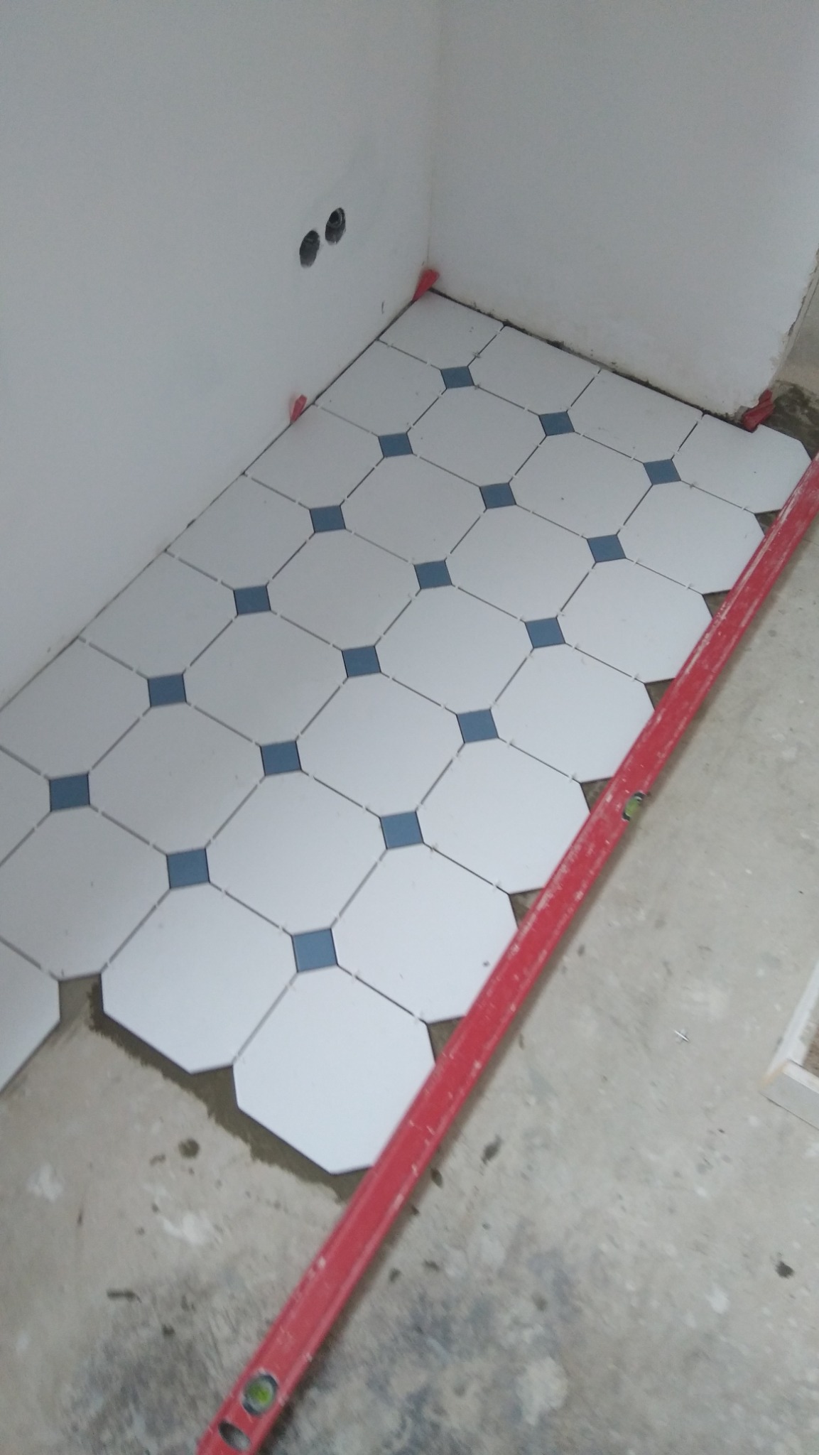 Układanie białych, ośmiokątnych płytek z niebieskim akcentem na podłodze w rogu pomieszczenia. Widoczna poziomica i krzyżyki dystansowe.