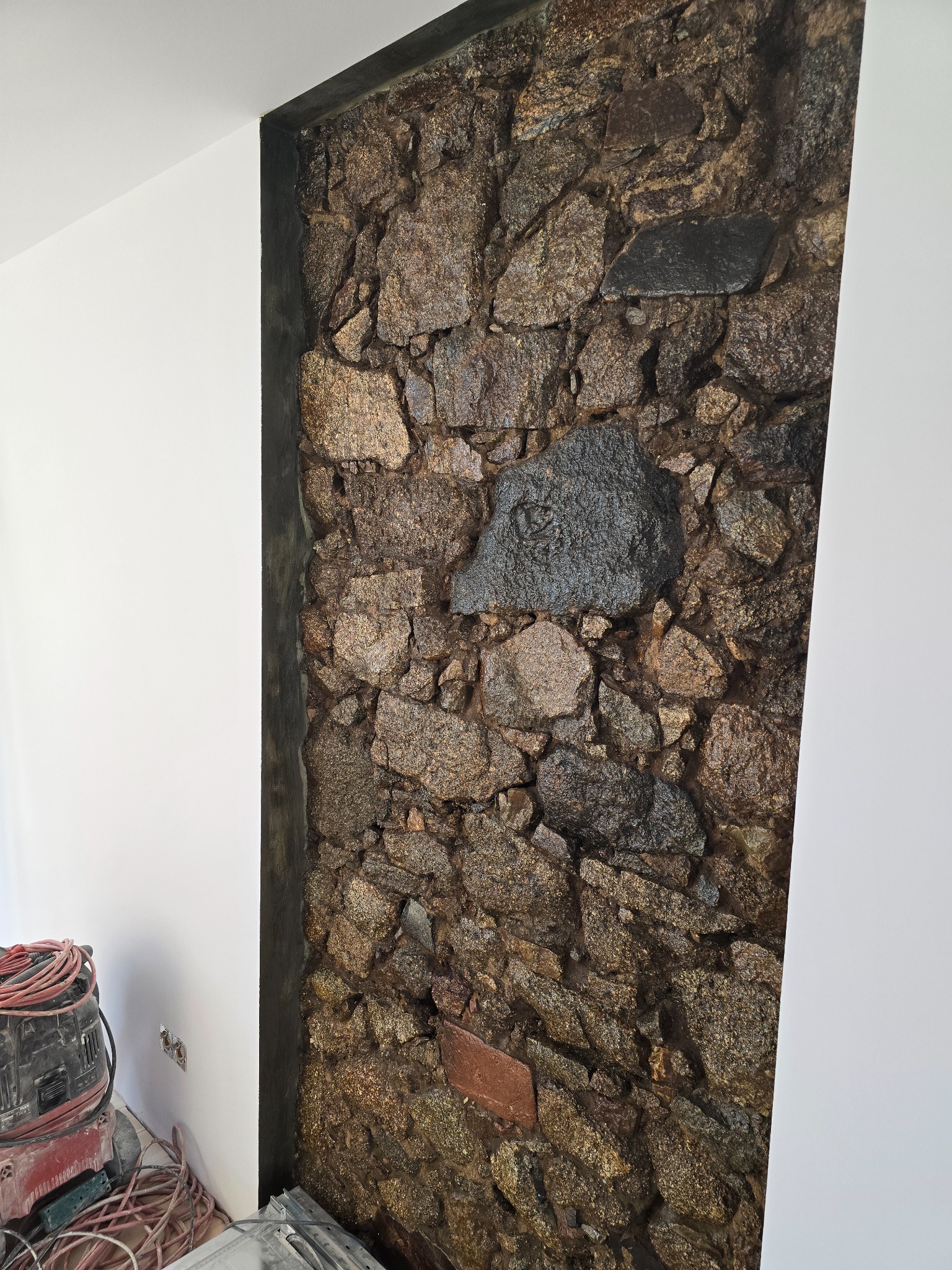 Dekoracyjna ściana z kamienia w aranżacji wnętrza, z widocznym fragmentem białej ściany i narzędziami budowlanymi. Wykończenie z surowym, naturalnym akcentem.