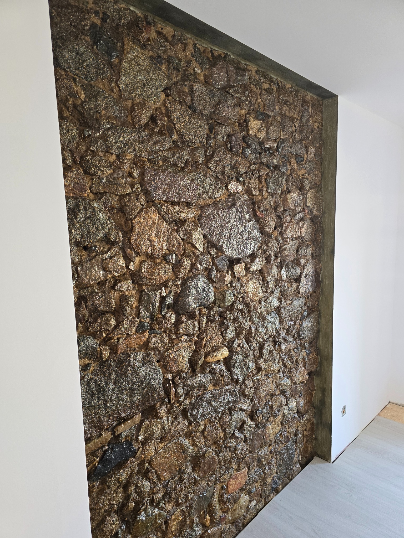 Ściana z kamienia dekoracyjnego w wykończonym wnętrzu, z fragmentem białej ściany i jasną podłogą. Widoczne nieregularne kształty i faktura kamieni.