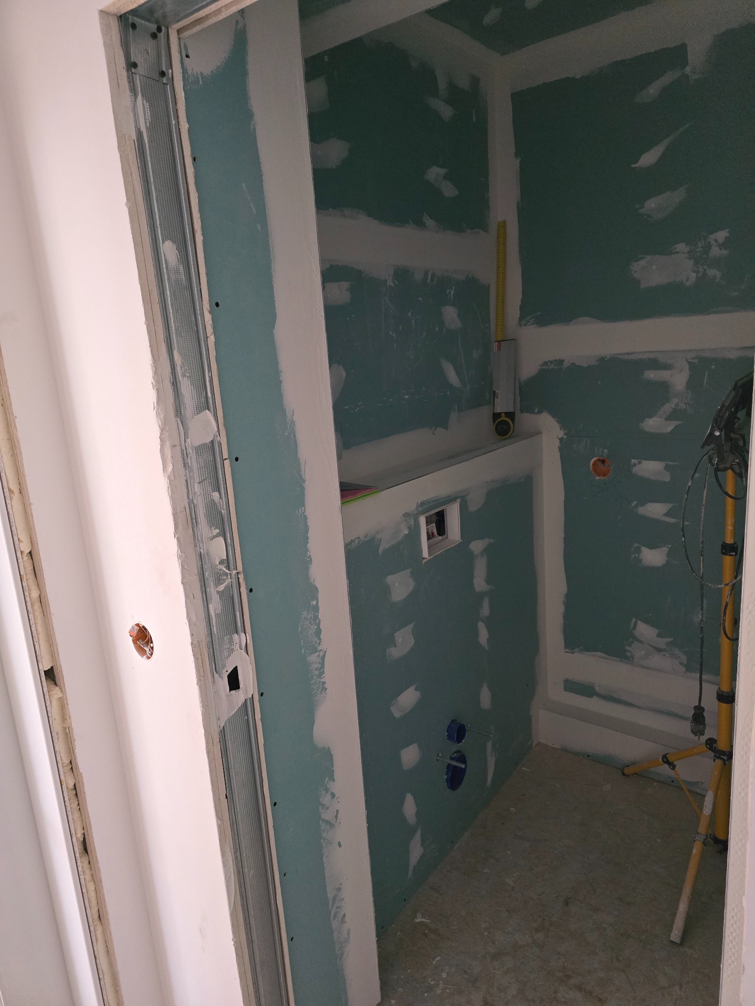 Łazienka w trakcie remontu: ściany z płyt gipsowo-kartonowych, widoczne instalacje, metalowe profile przy drzwiach, lampa budowlana. Etap prac wykończeniowych.