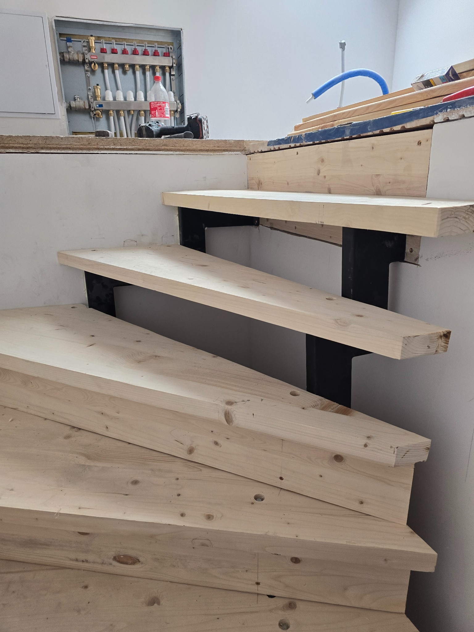 Wnętrze w trakcie remontu: drewniane schody z czarnymi wspornikami, instalacja hydrauliczna na ścianie. Widoczne narzędzia i materiały budowlane.