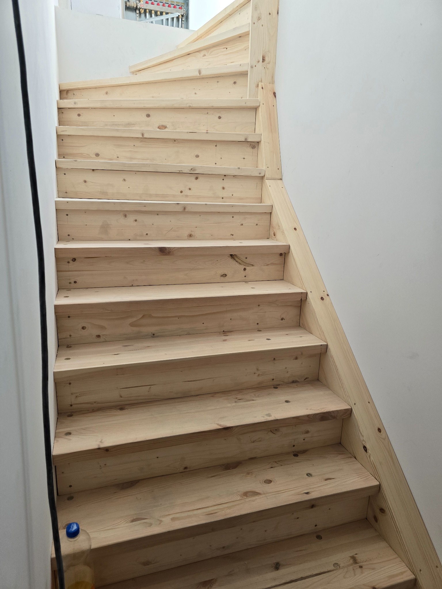 Jasne, drewniane schody wewnętrzne, świeżo po montażu, widoczne sęki i naturalna struktura drewna. Białe ściany i fragment instalacji na górze kadru.