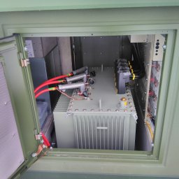 Przedsiębiorstwo Elektrotechniczne Konstelekt - Otwarta szafa elektryczna w kolorze khaki z widocznym transformatorem, przewodami w czerwonej izolacji i elementami instalacji elektrycznej.