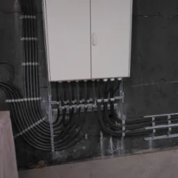 Biała skrzynka rozdzielcza na szorstkiej, ciemnej ścianie, z licznymi czarnymi kablami w metalowych uchwytach, prowadzącymi do i od skrzynki.