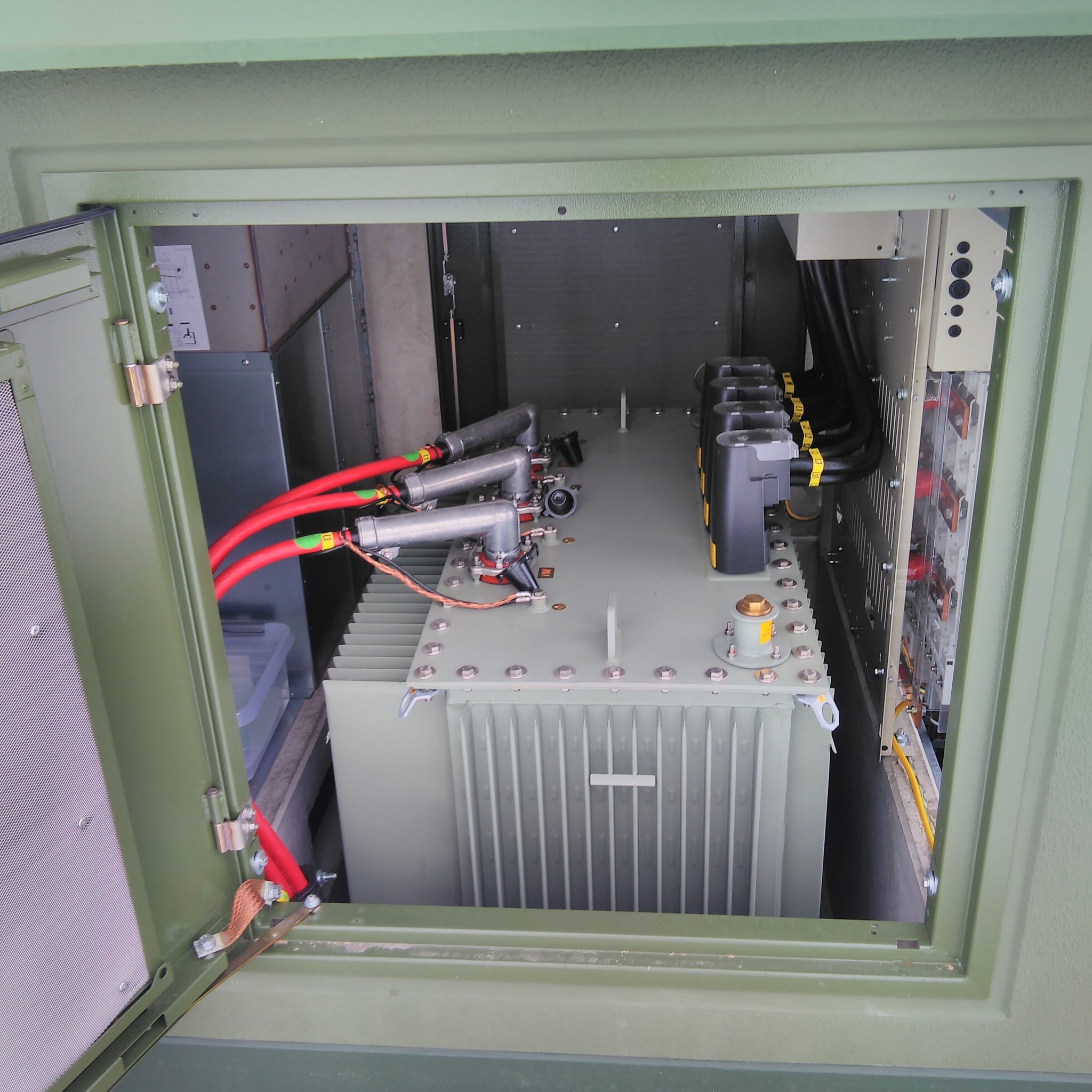 Otwarta szafa elektryczna w kolorze khaki z widocznym transformatorem, przewodami w czerwonej izolacji i elementami instalacji elektrycznej.