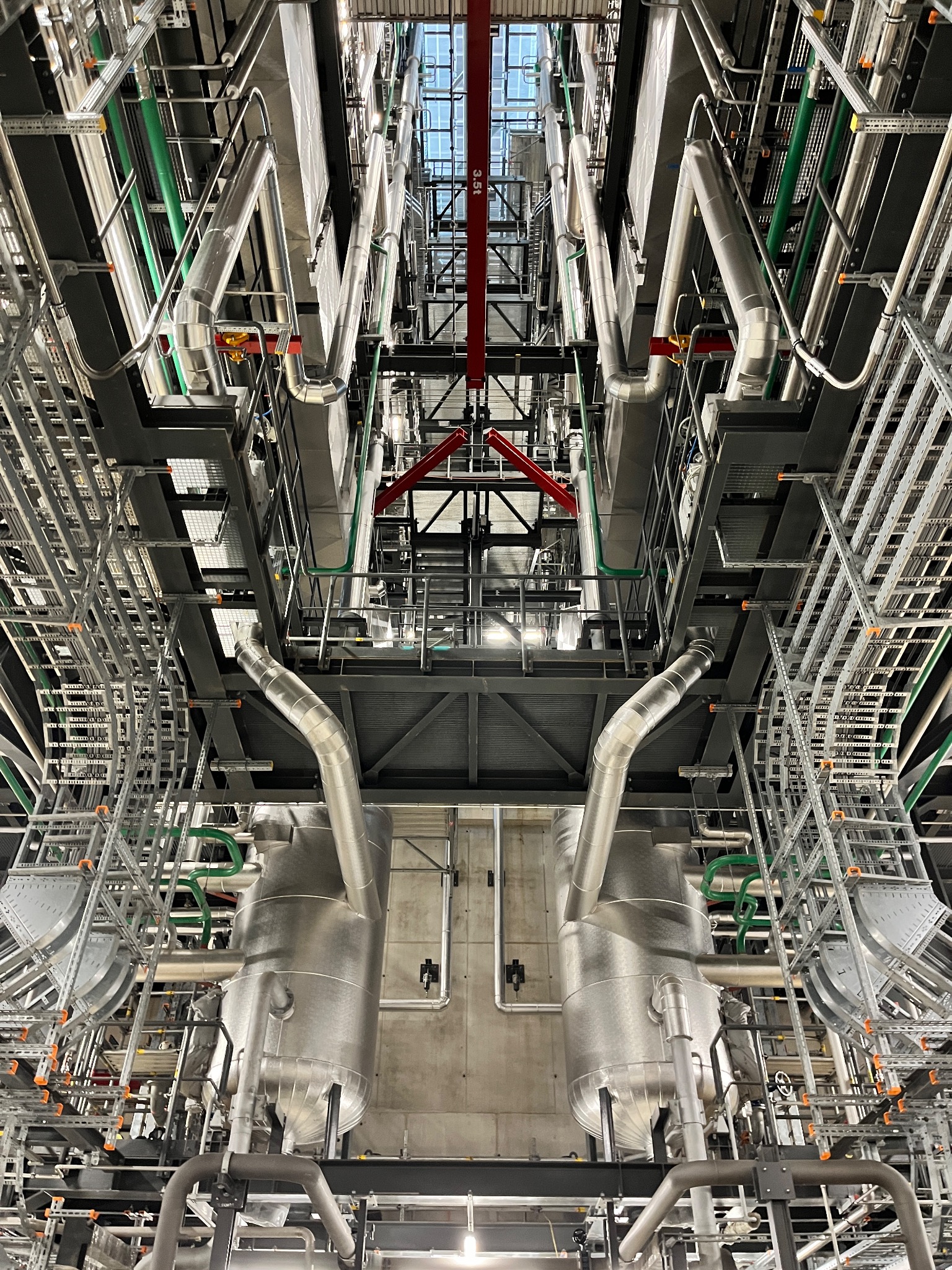 Widok z dołu na skomplikowaną sieć rur, kabli i konstrukcji wsporczych w hali przemysłowej, z elementami w kolorze srebrnym, zielonym i czerwonym, oświetlonych światłem dziennym wpadającym przez...