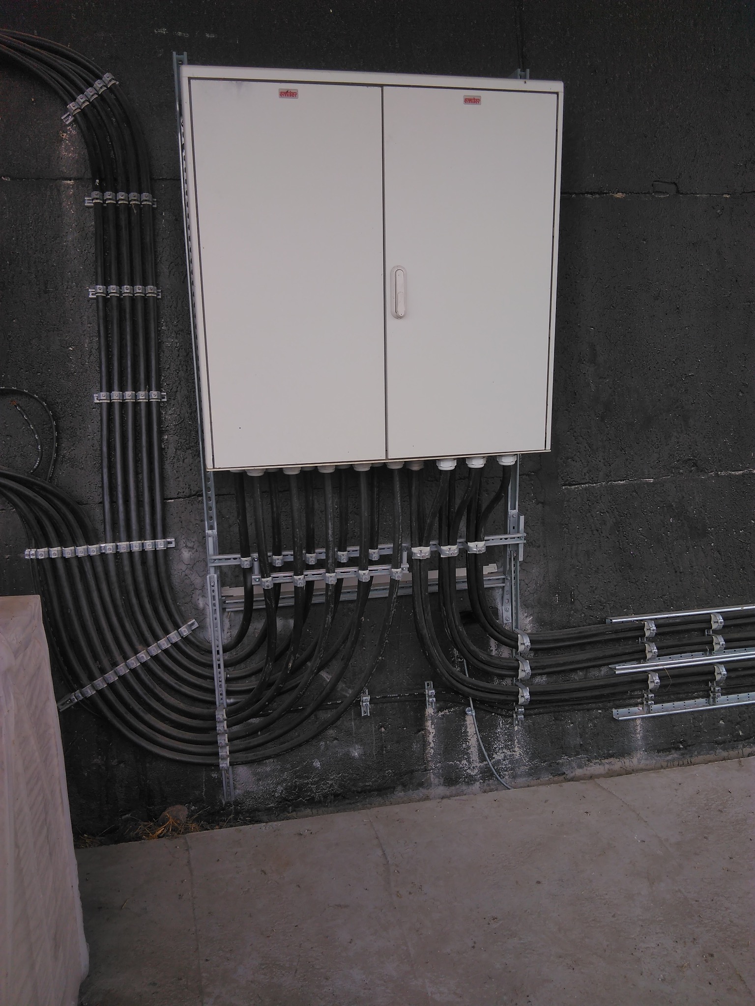 Biała skrzynka rozdzielcza na szorstkiej, ciemnej ścianie, z licznymi czarnymi kablami w metalowych uchwytach, prowadzącymi do i od skrzynki.