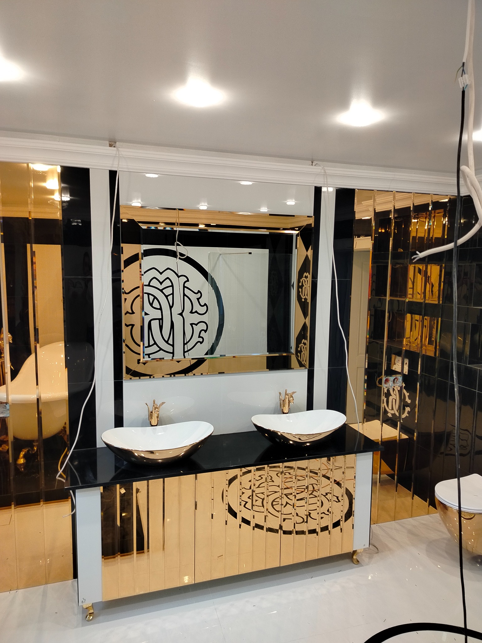 Luksusowa łazienka z dwoma umywalkami na czarnym blacie, złote płytki na ścianach i szafce, lustro z dekoracyjnym motywem. Widoczne elementy instalacji elektrycznej.