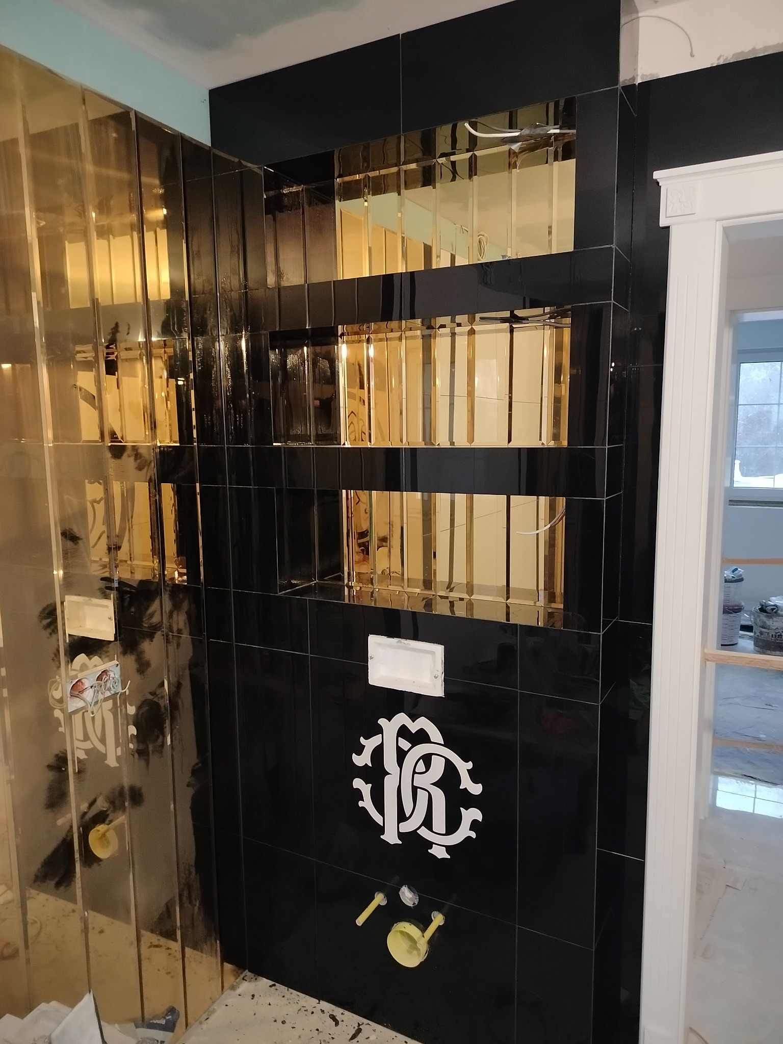 Ściana łazienki wykończona czarnymi i złotymi płytkami, z dekoracyjnym wzorem. Widoczne elementy instalacji i fragment drzwi. Nietypowe połączenie kolorów.