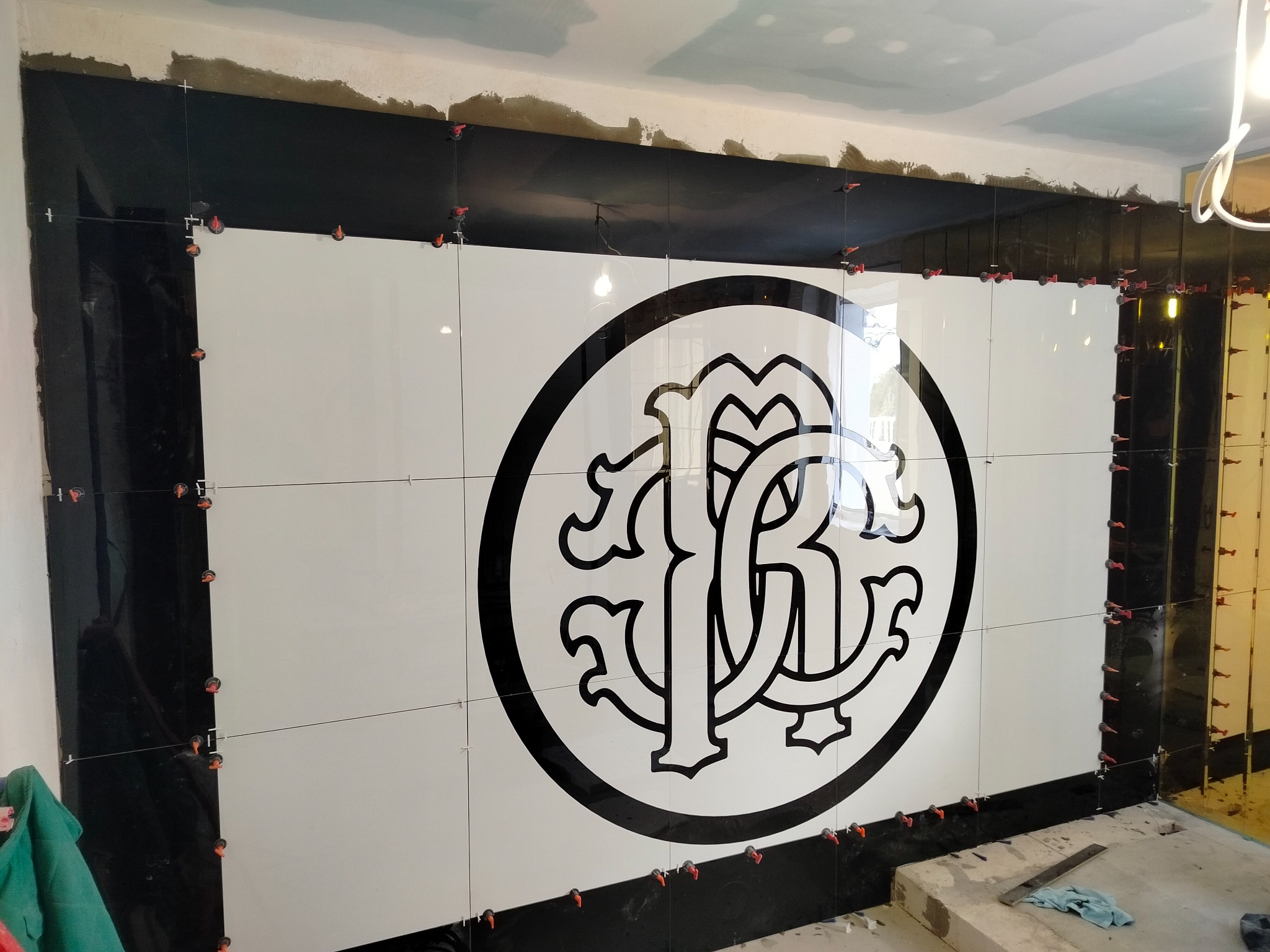 Czarno-biała ściana z błyszczących płytek z logo, w trakcie montażu, z użyciem poziomicy i krzyżyków dystansowych. Wykończenie łazienki w nowoczesnym stylu.