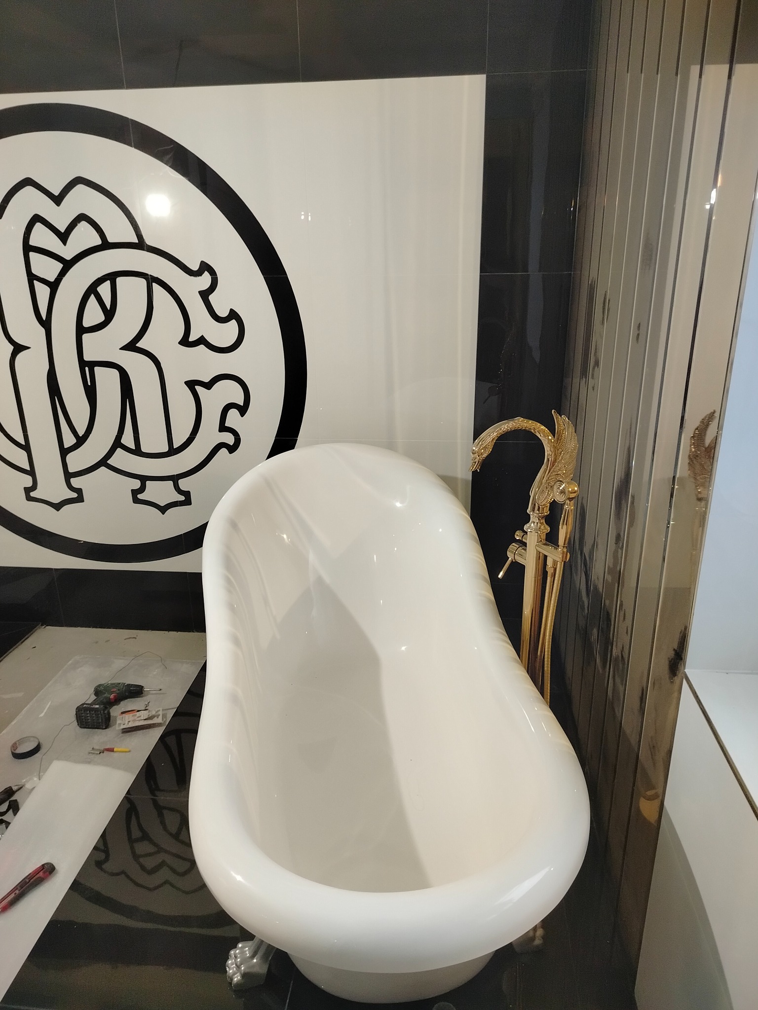 Biała wanna wolnostojąca z ozdobną złotą armaturą na tle czarno-białych płytek z wzorem. Na podłodze widoczne narzędzia glazurnicze.